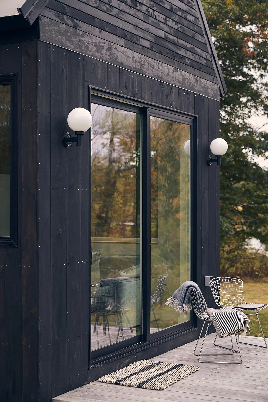 Exterior view of a modern black wooden house with glass sliding doors, outdoor seating area with two white wire chairs, blankets, a rug, and surrounded by trees.