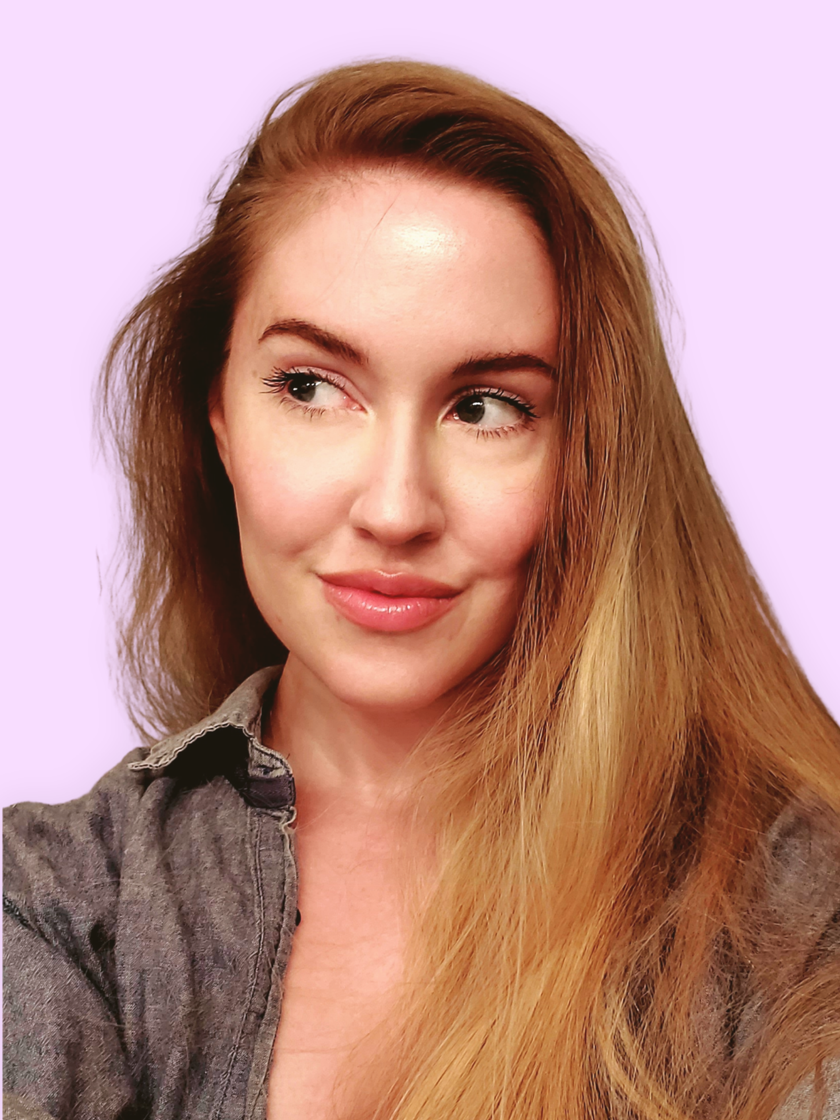 Close-up of a woman with long hair, wearing a grey denim shirt, smiling softly against a light purple background.