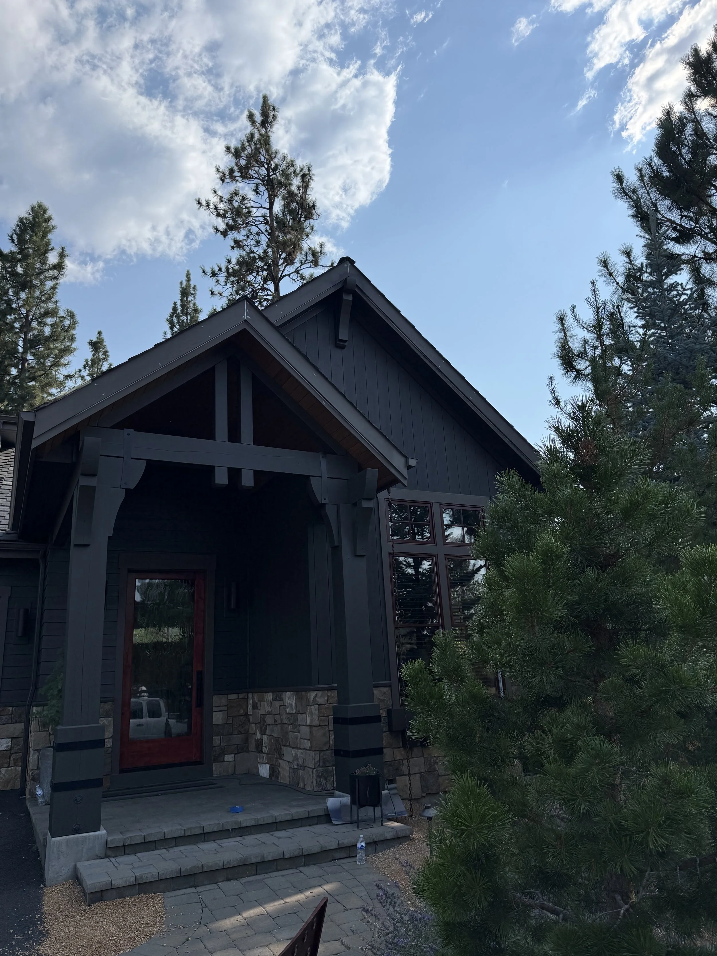 The image shows the exterior of a modern house with a dark-colored facade, stone accents, and a large window. There is a small porch with steps and a paved area in front, surrounded by tall pine trees under a partly cloudy sky.