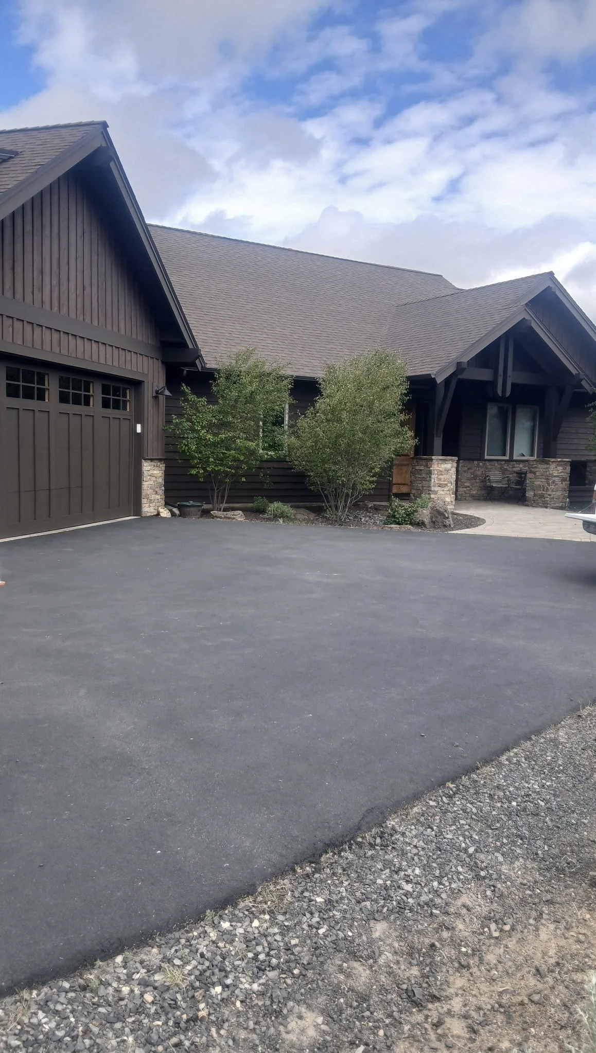 Newly paved black asphalt driveway in front of a house with dark brown wooden siding, stone accents, and a garage door on the left. There are two small green trees and some shrubs near the house. The sky is partly cloudy with patches of blue.
