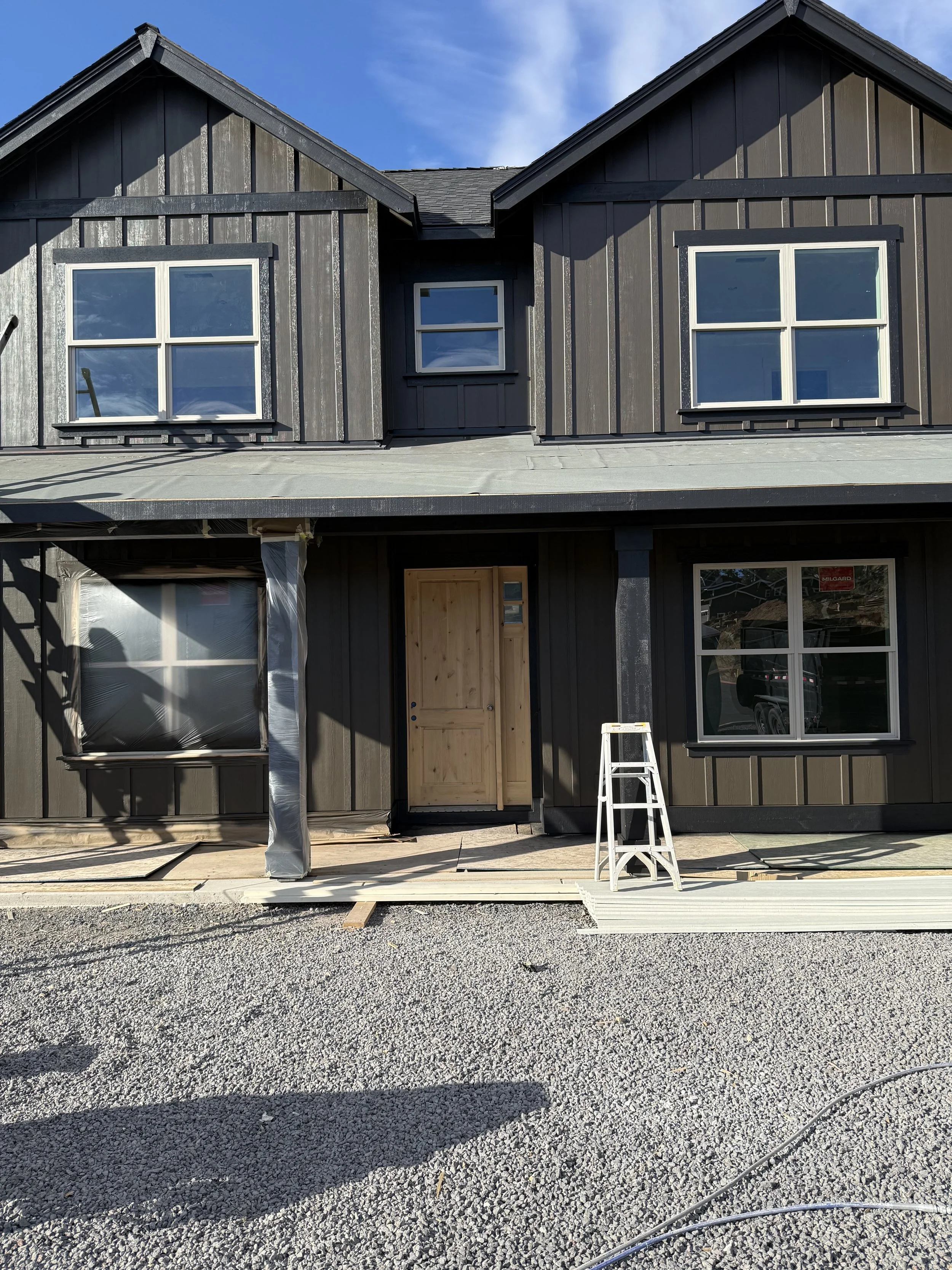 Construction site of a modern house with dark wooden exterior, four white-framed windows on the upper floor, and an unfinished entryway on the lower floor with a wooden door frame. A small ladder and construction materials are visible in the foregrou