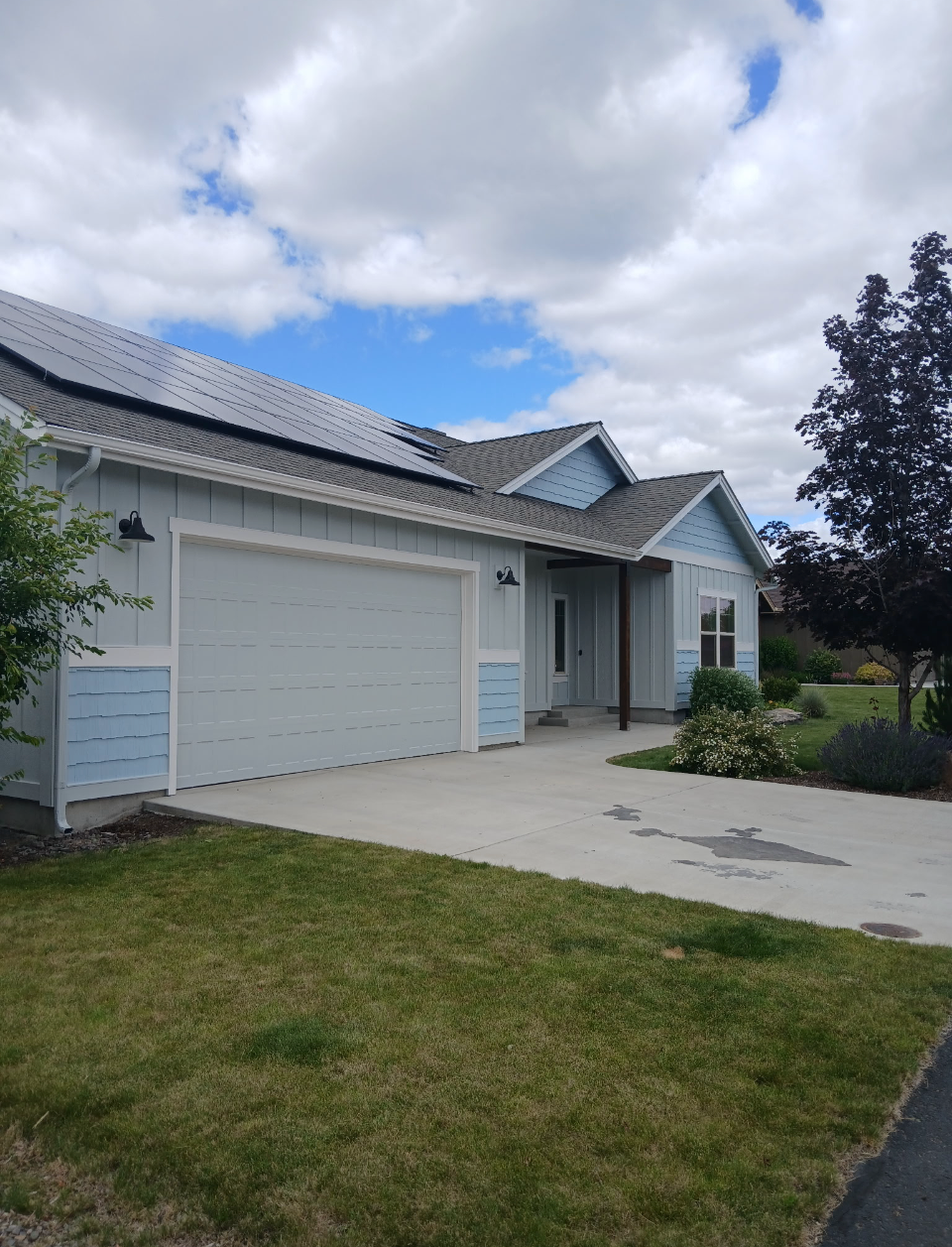 Front view of a light blue house with a garage, solar panels on the roof, and a concrete driveway, surrounded by lawn and bushes under a partly cloudy sky.