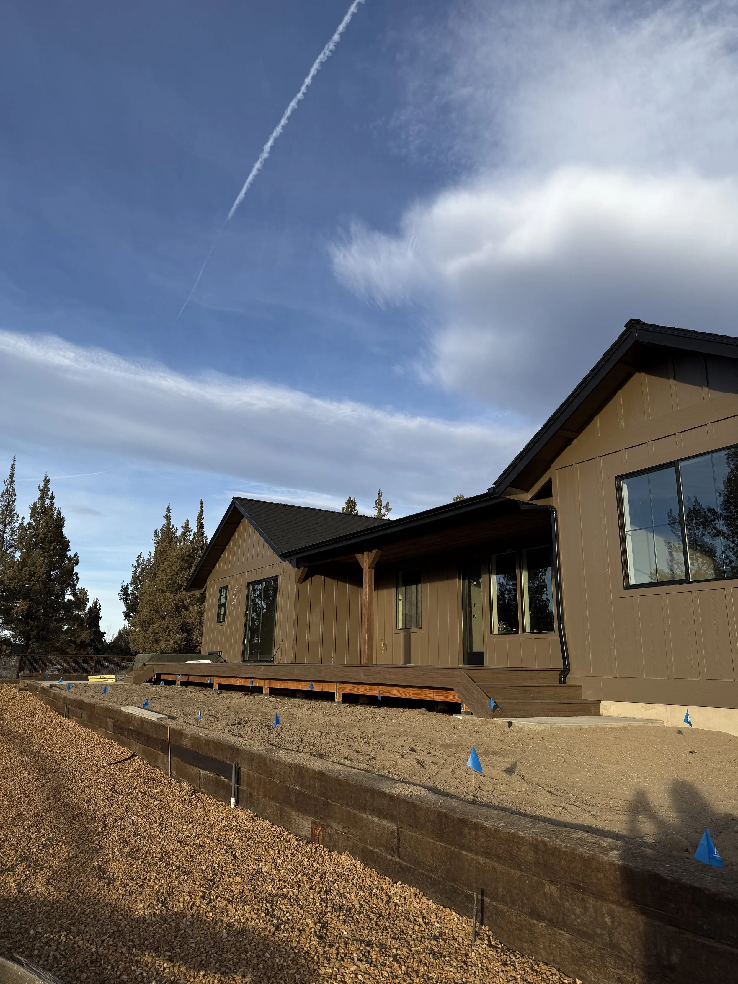 A house under construction with a raised wooden deck, beige exterior, large windows, surrounded by a dirt yard with small blue construction flags and a backdrop of tall trees and a partly cloudy sky.