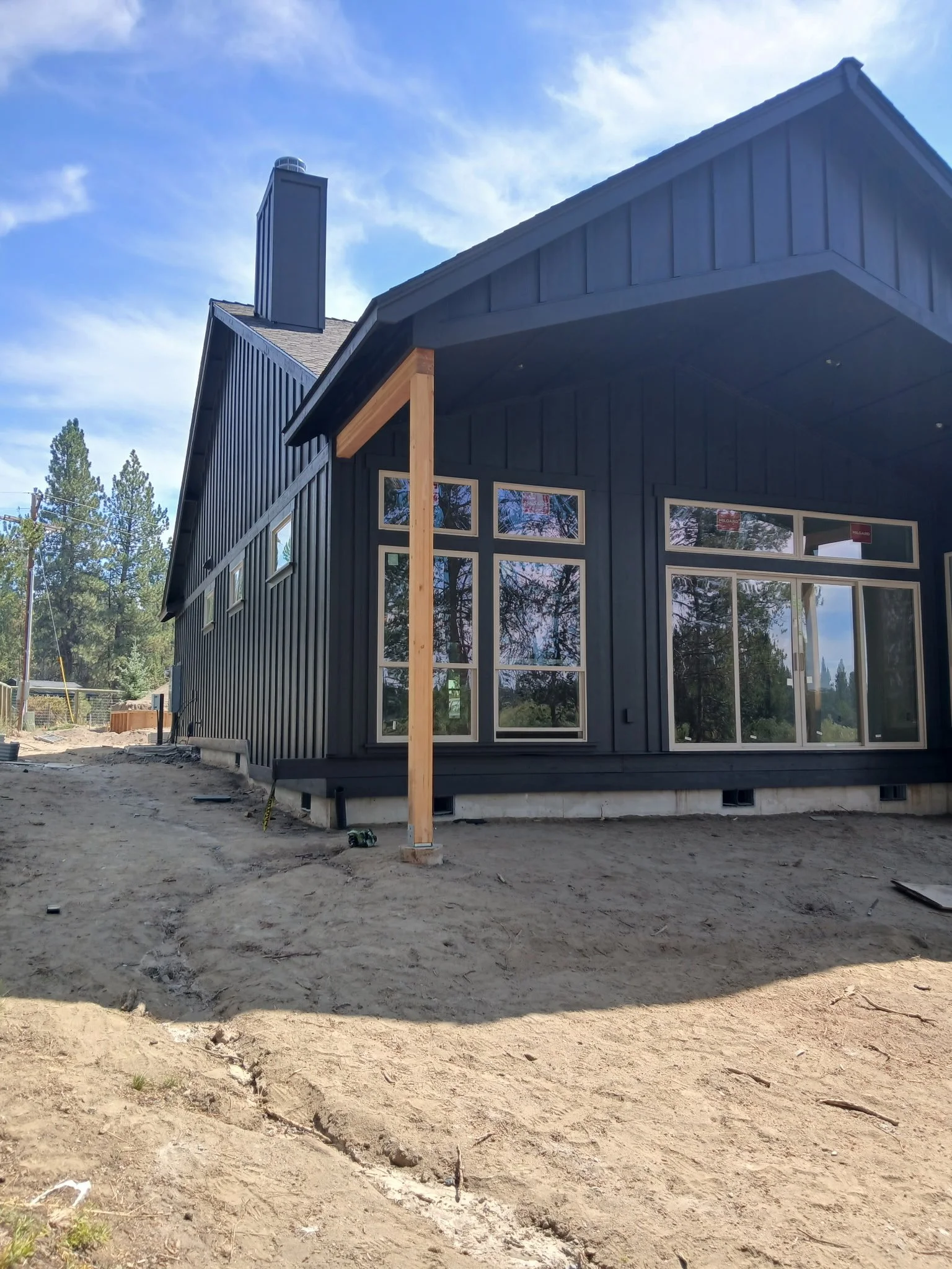 A two-story house under construction with dark exterior siding, multiple large windows, a chimney, and a front porch supported by a wooden post, set in a natural outdoor environment with trees in the background.