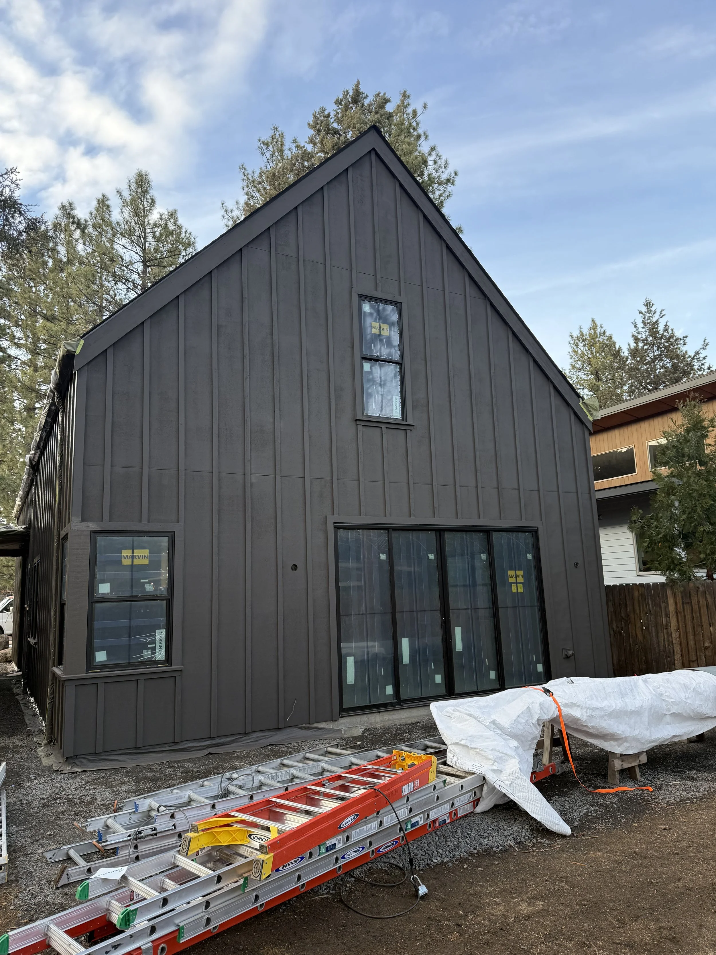 A two-story house under construction with black vertical siding, a high peaked roof, and multiple windows. Construction tools and materials, including ladders and plastic coverings, are on the ground in front of the house. Trees and a partly cloudy s