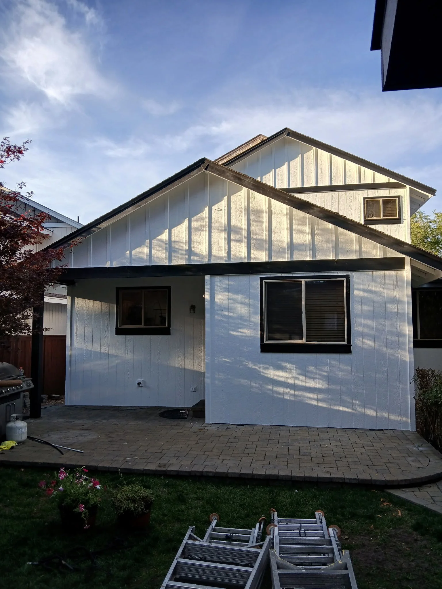A backyard patio with pavers, green grass, potted plants, and a ladder. A two-story white house with black window frames, a small window, and a slanted roof is in the background. Part of a neighboring house is visible, and the sky is partly cloudy.