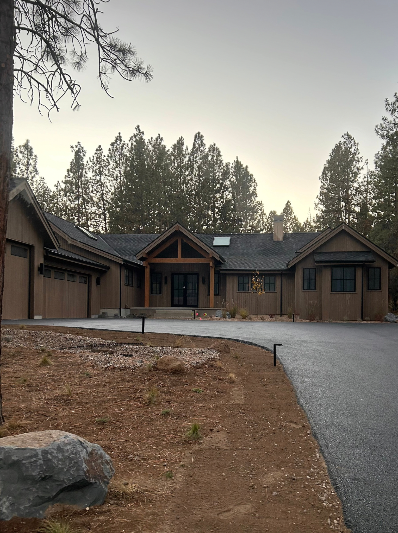 View of a modern house with a dark roof, large windows, and a front porch, surrounded by a dirt yard with small plants and rocks, against a backdrop of tall pine trees and a cloudy sky.