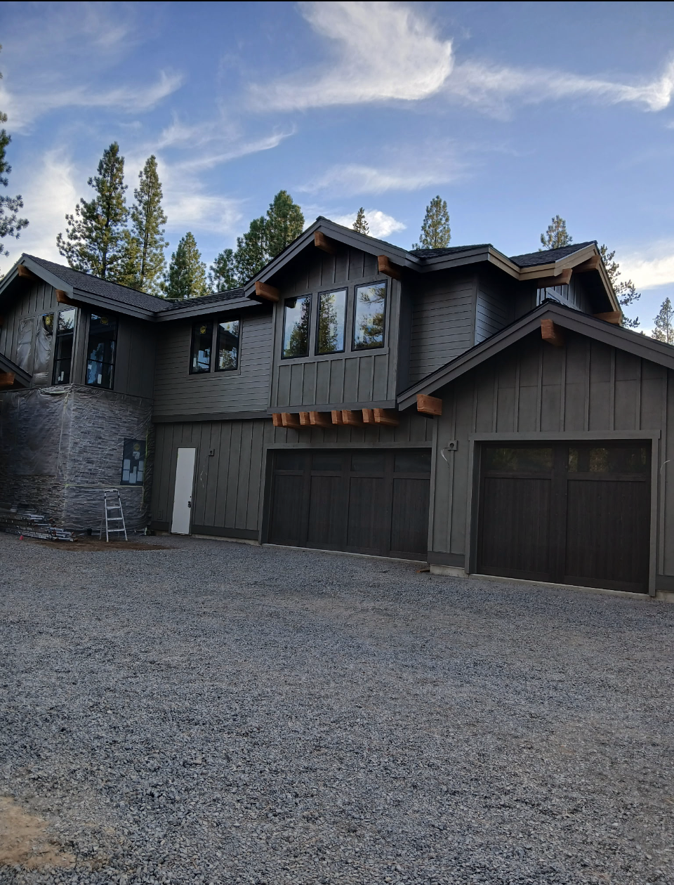 Newly constructed modern two-story house with black exterior, multiple large windows, and attached garage, surrounded by tall pine trees and a gravel driveway under a partly cloudy sky.