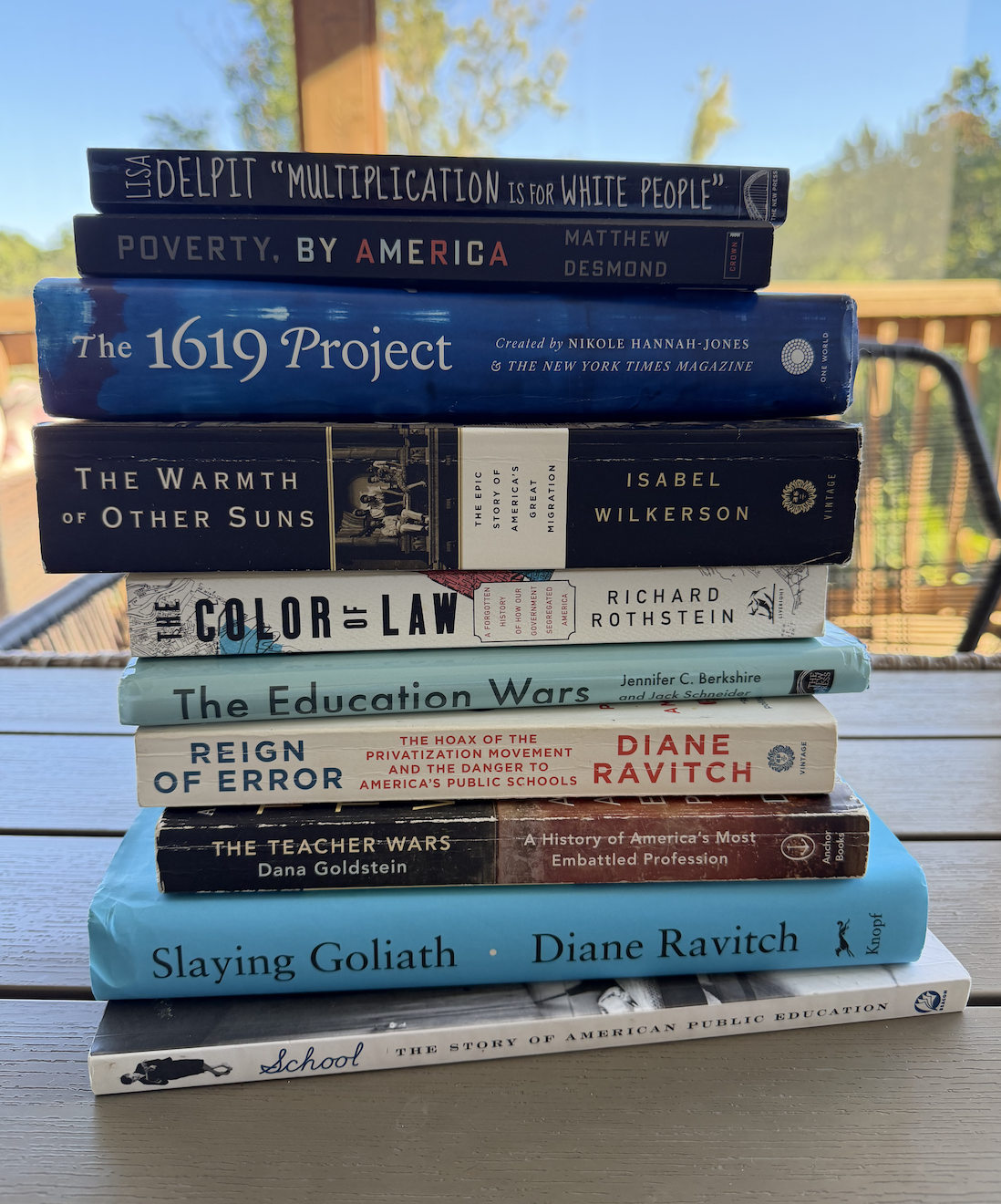 Stack of ten books on a wooden table outdoors, with trees and a blue sky in the background.