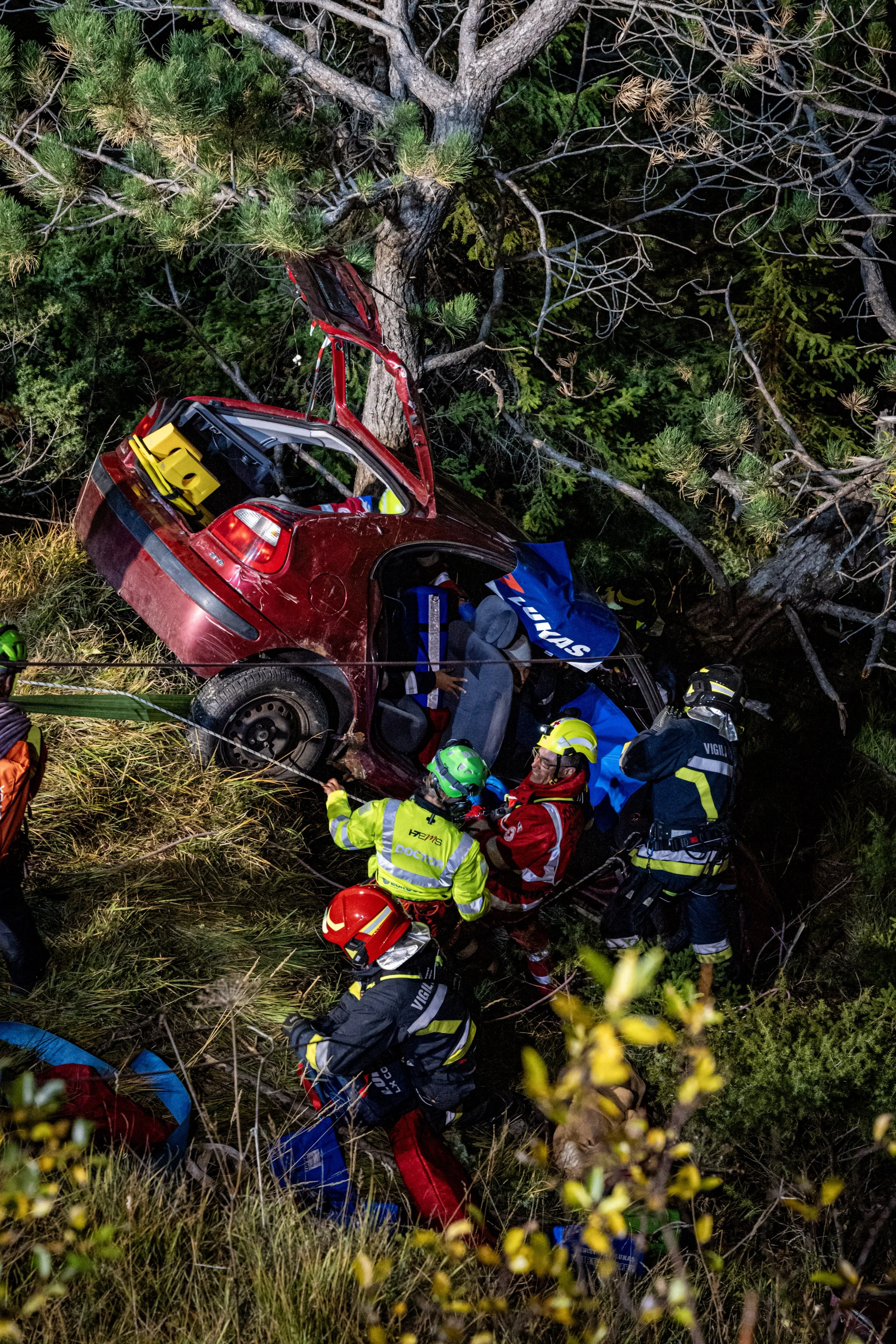 Emergency rescue personnel attend to a car crash scene involving a red vehicle that has collided with a tree in a forested area at night. The scene is illuminated, and rescue workers are wearing helmets and reflective gear, working around the car ami