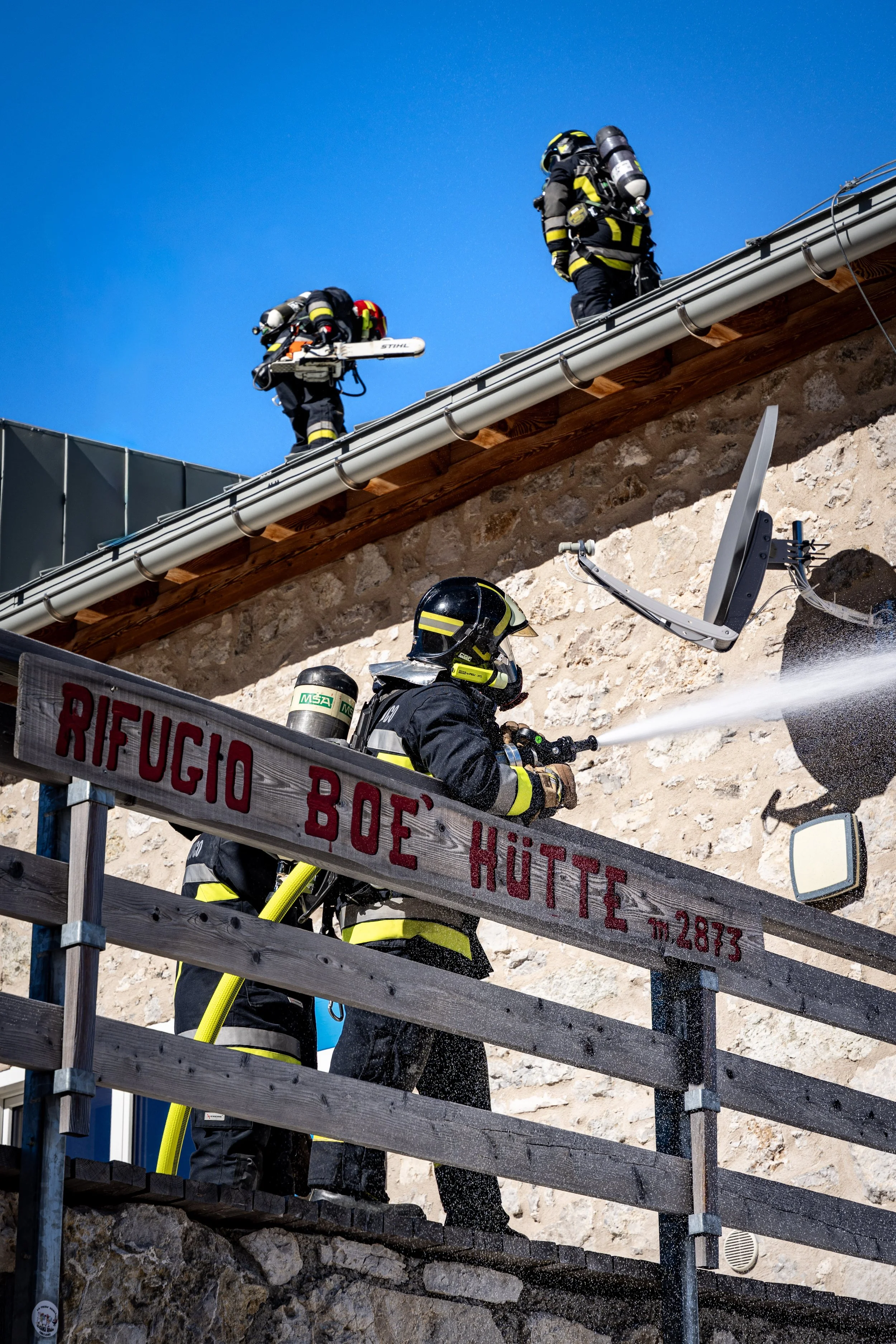 Firefighters in protective gear on a ladder cleaning a building's exterior wall with a hose, with a blue sky background.