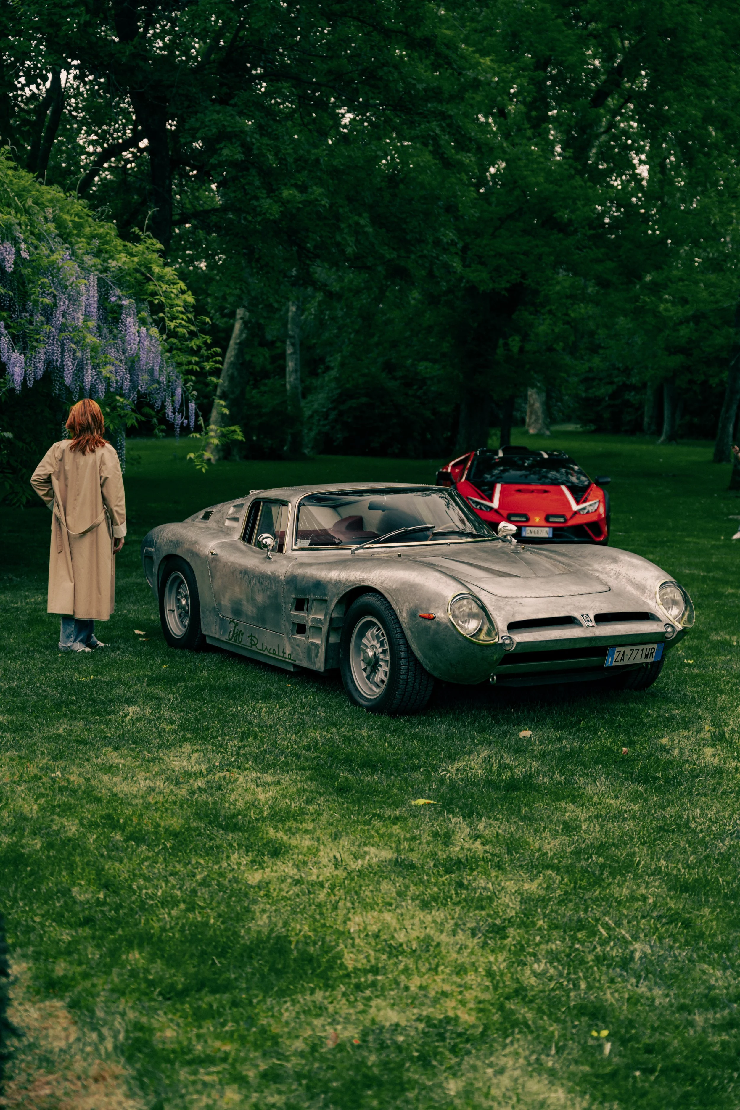 A woman with red hair wearing a beige trench coat standing on a grassy area, looking at two cars: an old silver sports car in the foreground and a modern red sports car in the background, surrounded by lush green trees.