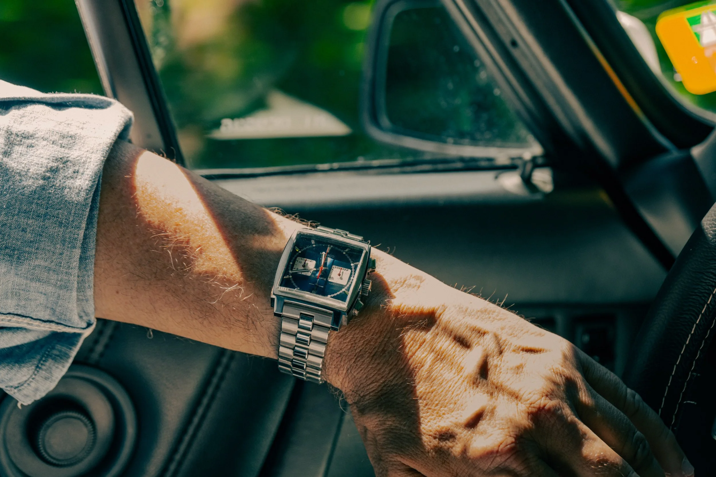 A person's arm resting on a car's dashboard, wearing a silver wristwatch with a blue dial, with the inside of the car and side mirror in the background.
