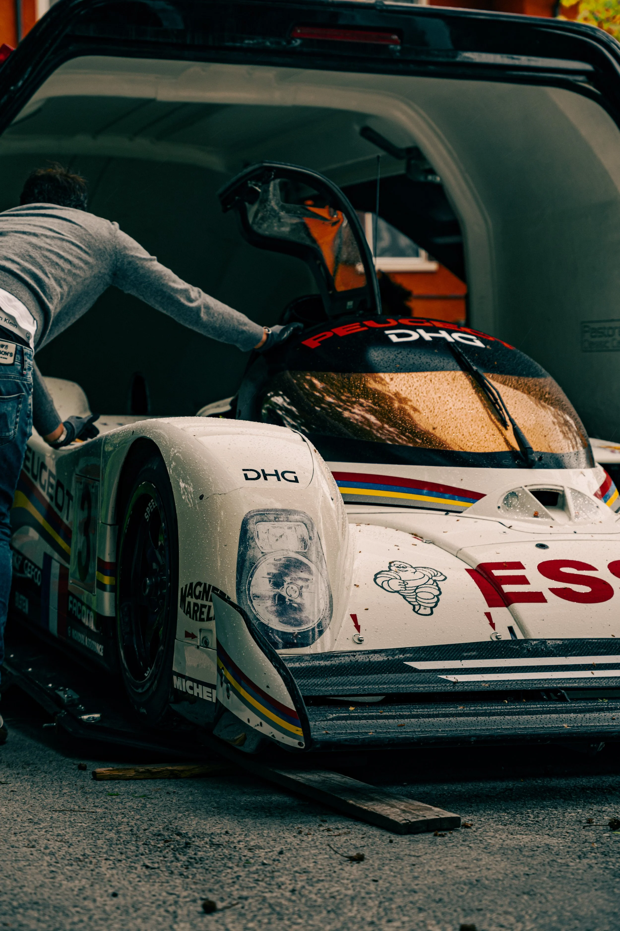 A racing car parked in a garage, with a mechanic reaching inside. The car has various sponsorship logos, including Michelin and Michelin Man, and is predominantly white with colorful stripes.