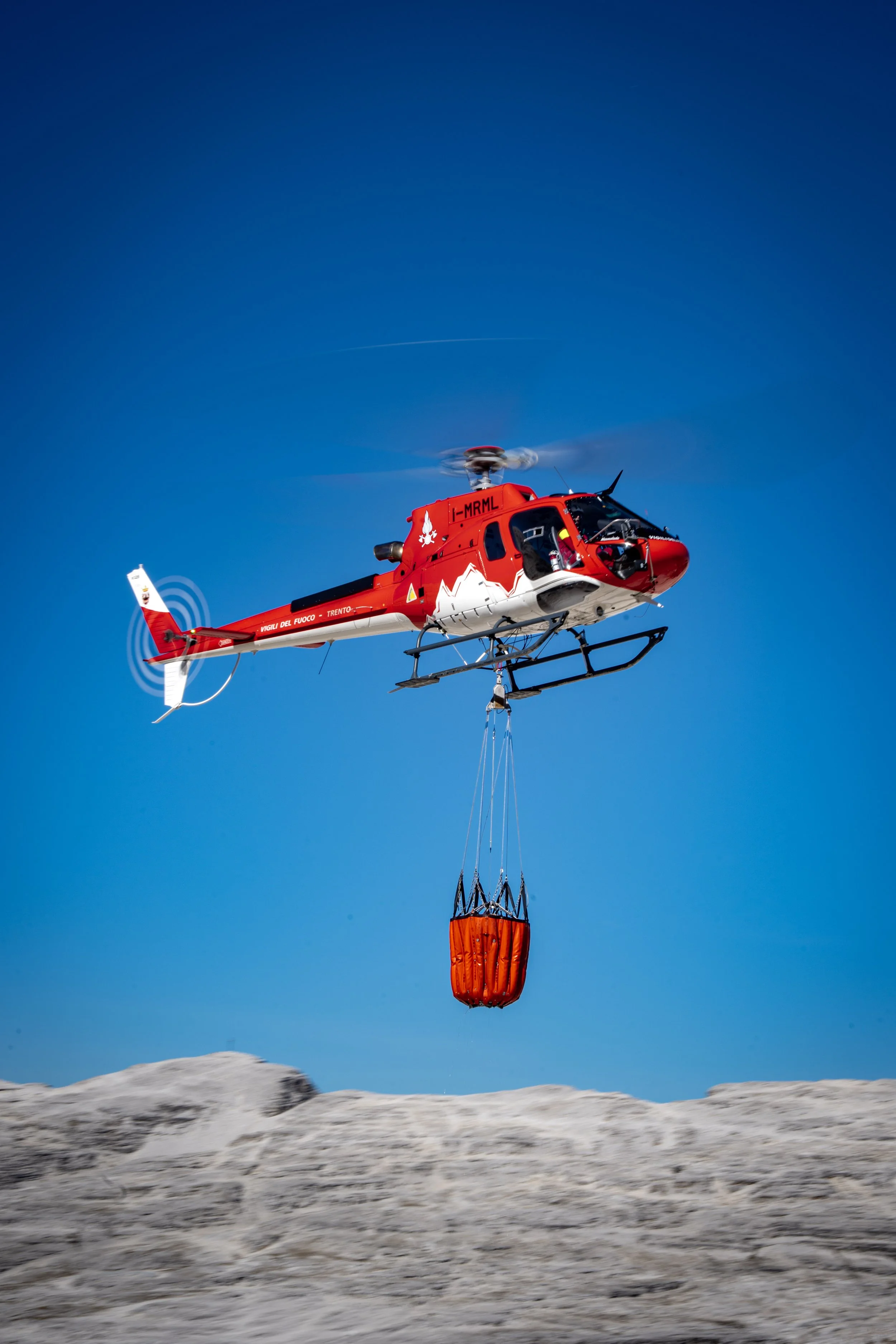A helicopter flying in the sky carrying a rescue basket with supplies, above a snowy mountainous landscape.