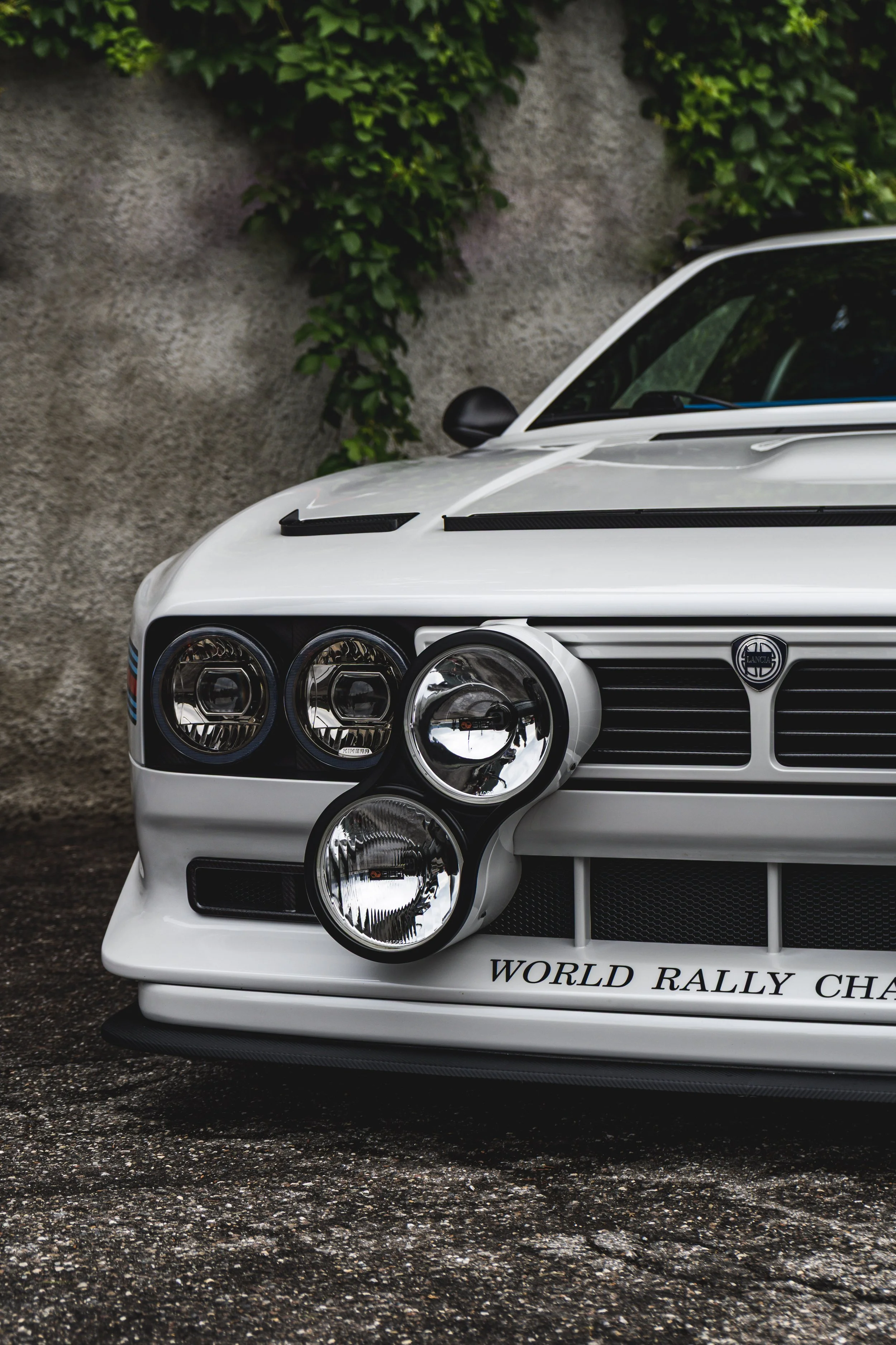 Close-up of a white rally car's front end showing detailed headlamps, grille with a Lancia badge, and partial text 'WORLD RALLY CH' on the front bumper. Background includes a textured concrete wall with green foliage.