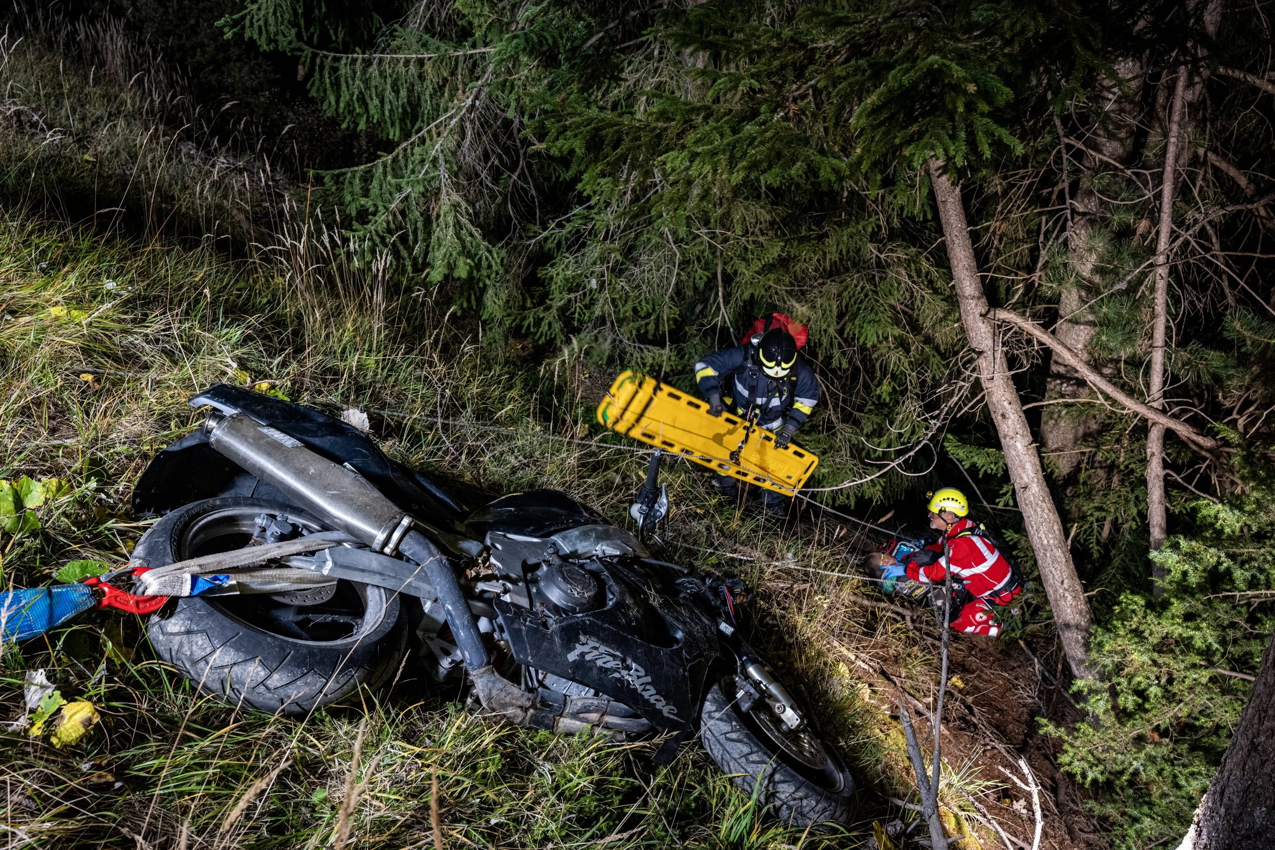 Emergency responders examining a crashed motorcycle in a wooded area at night, with emergency equipment and personnel present.