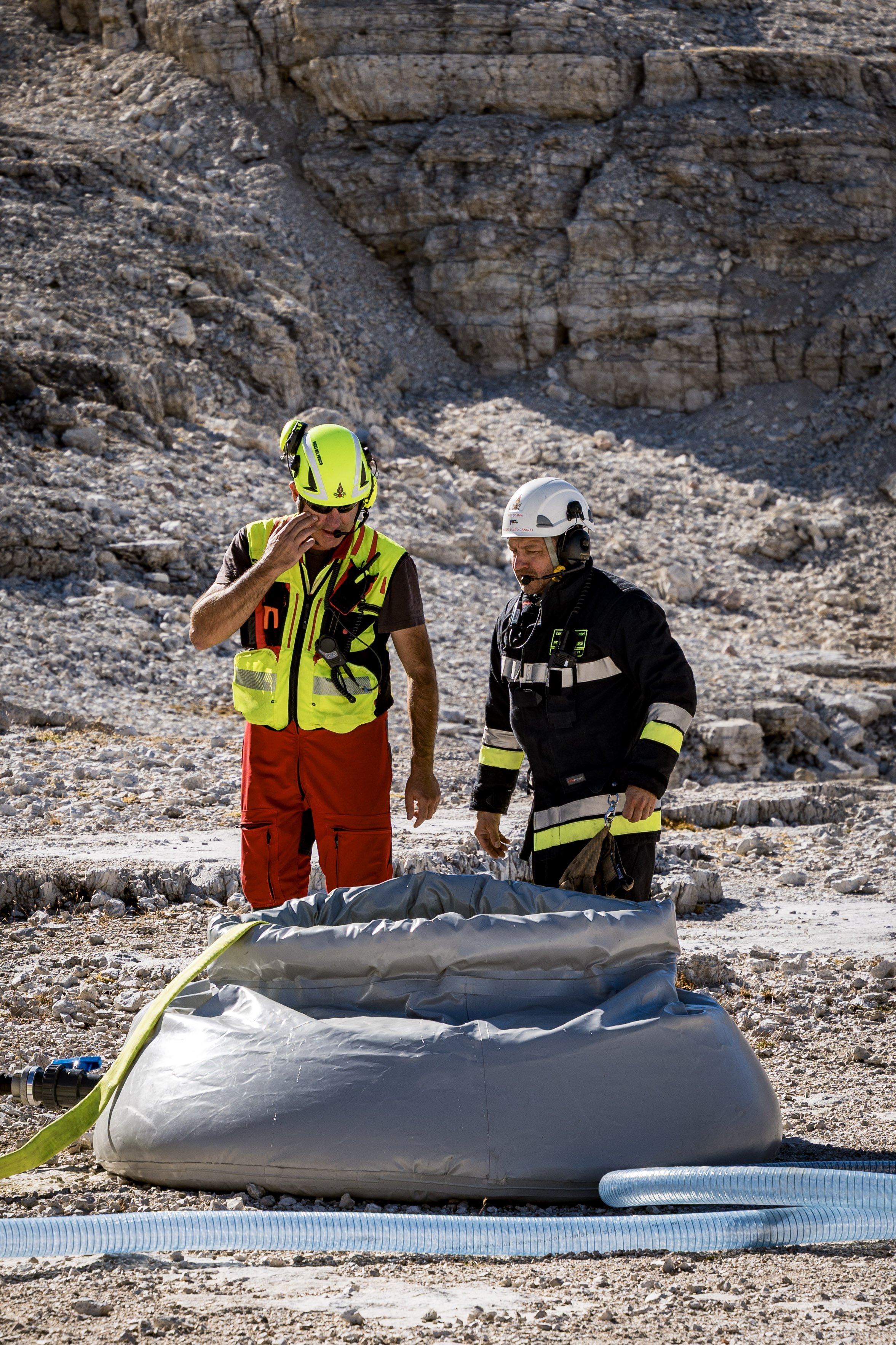 Two rescue workers in safety helmets and uniforms stand over a body covered with a tarp on rocky terrain with a rock cliff in the background. Equipment such as a large bag, hoses, and tools are nearby.