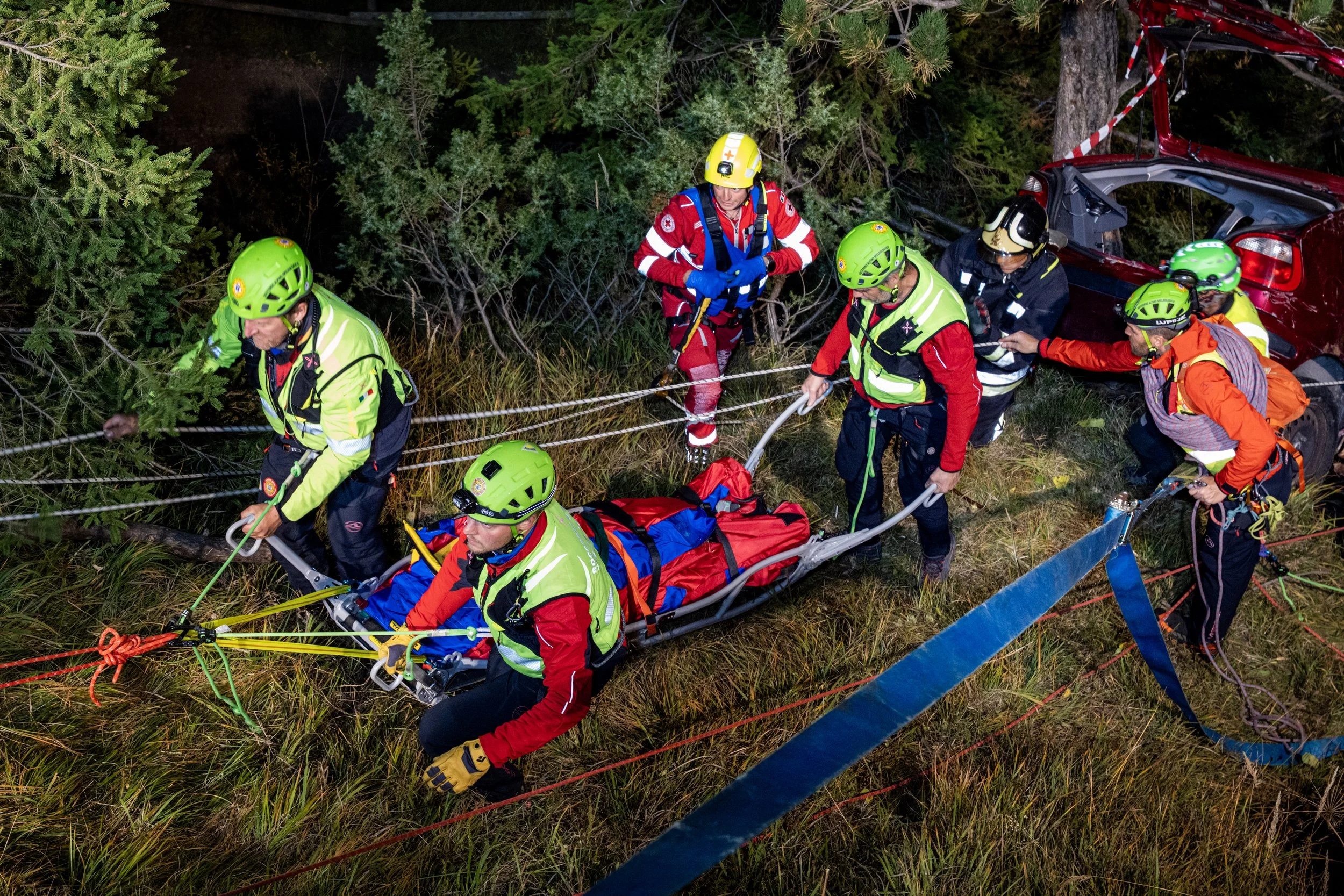 Emergency rescue team members in high-visibility gear and helmets are working together in a forest at night, positioning a stretcher with a person on it, near a damaged red car.