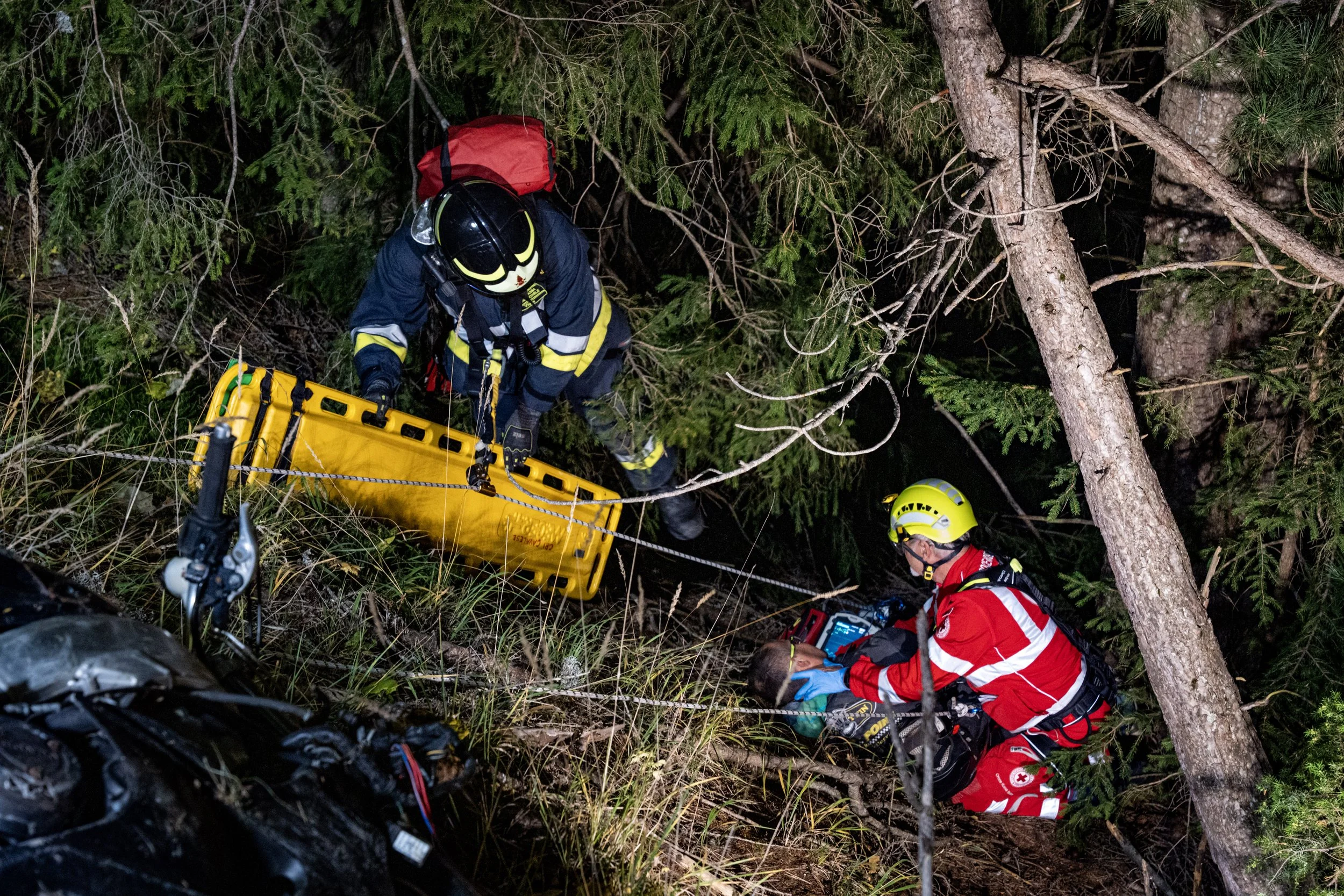 Rescue personnel in safety gear using a stretcher in a wooded area at night, presumably performing a rescue operation.