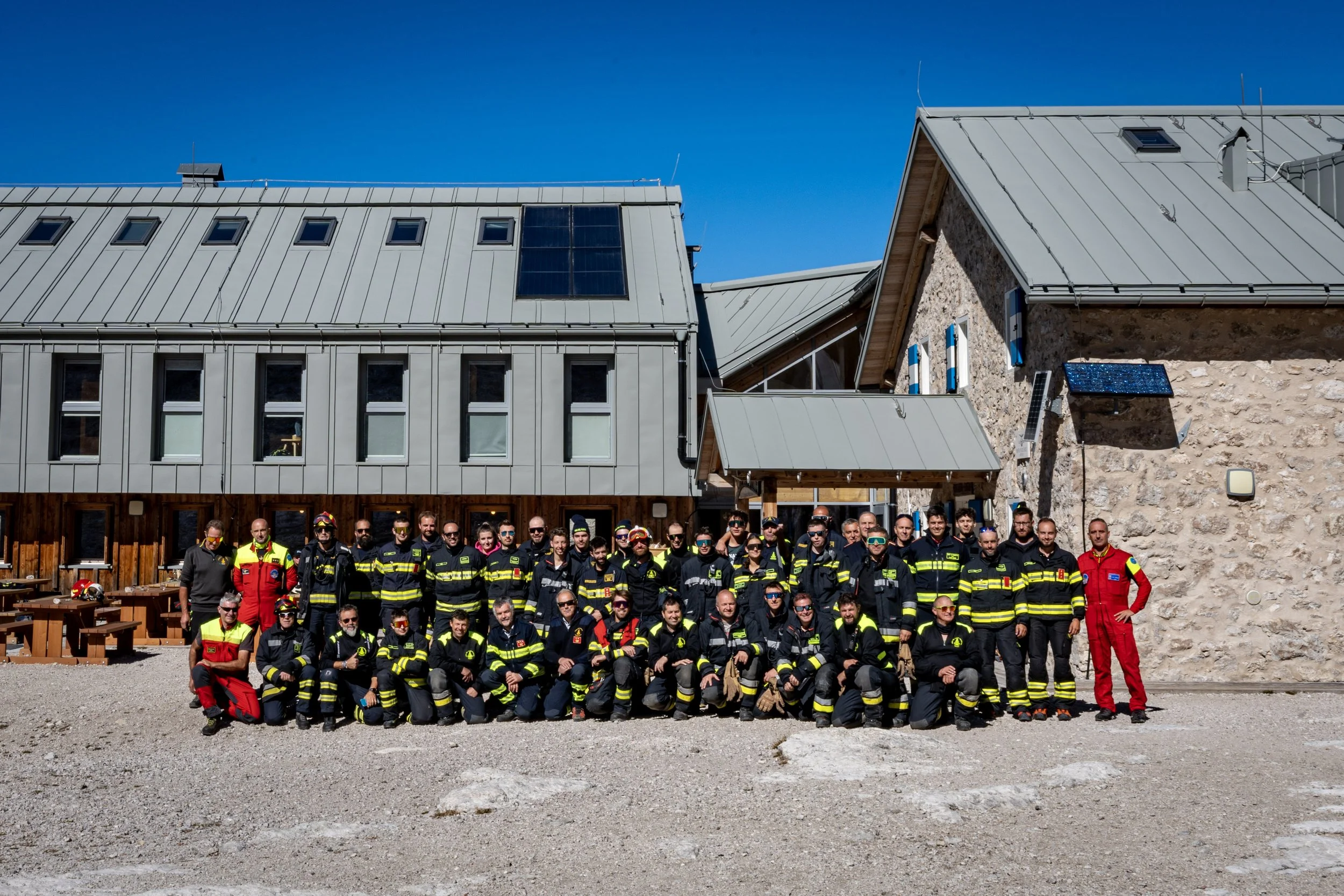 Group of emergency responders and firefighters standing together outdoors in front of a building with a metal roof and solar panels, under a clear blue sky.