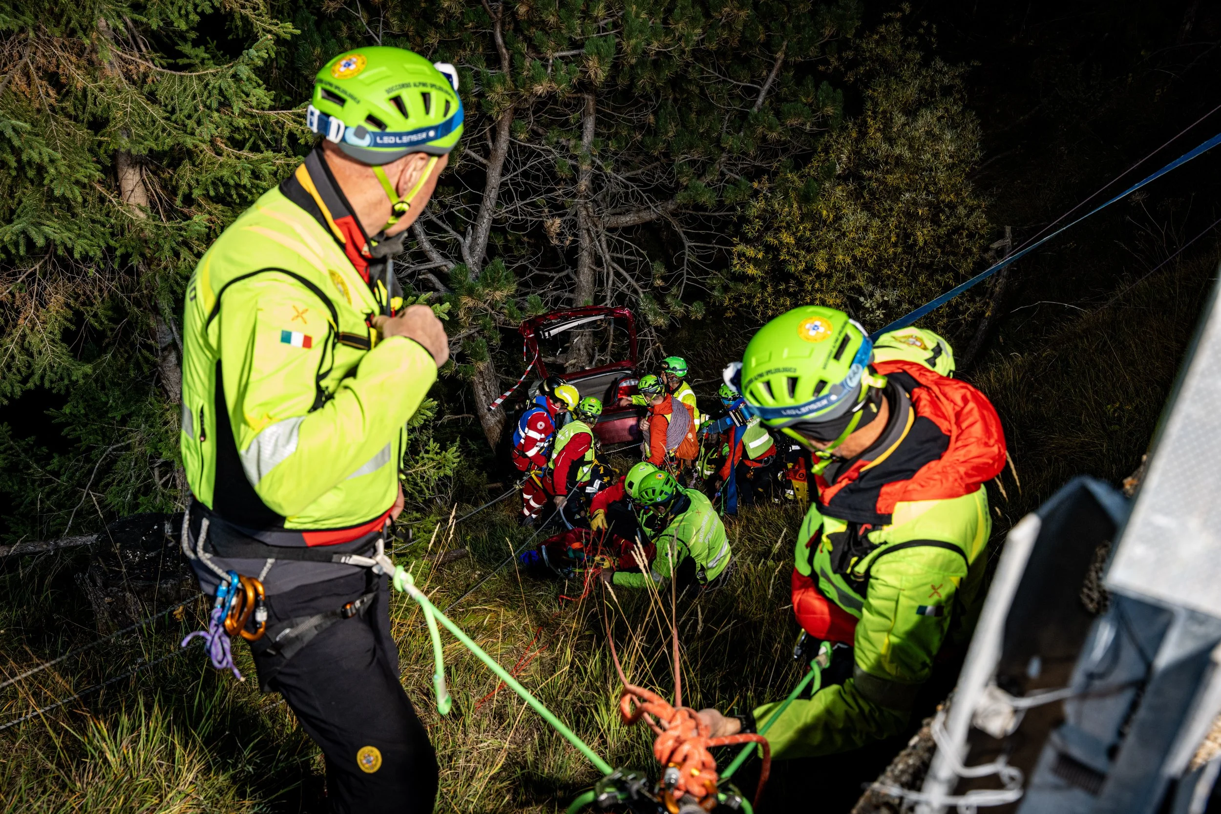 Emergency rescue team wearing bright yellow and orange gear, helmets, and harnesses, working together in a forested area to rescue a person from a vehicle that has gone off the road and is entangled among trees.