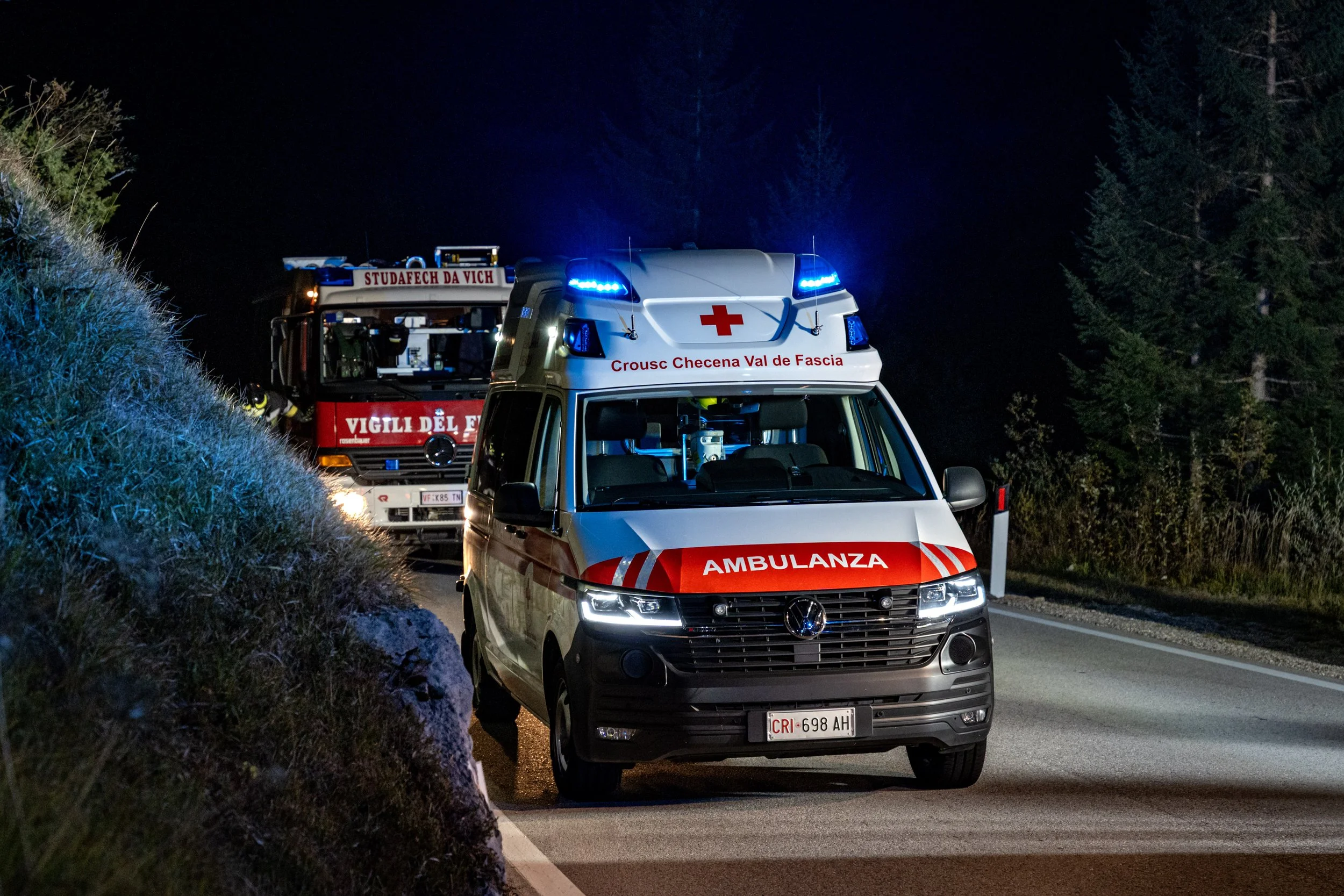 Emergency vehicles with flashing blue lights on a nighttime rural road, including an ambulance and a fire truck.