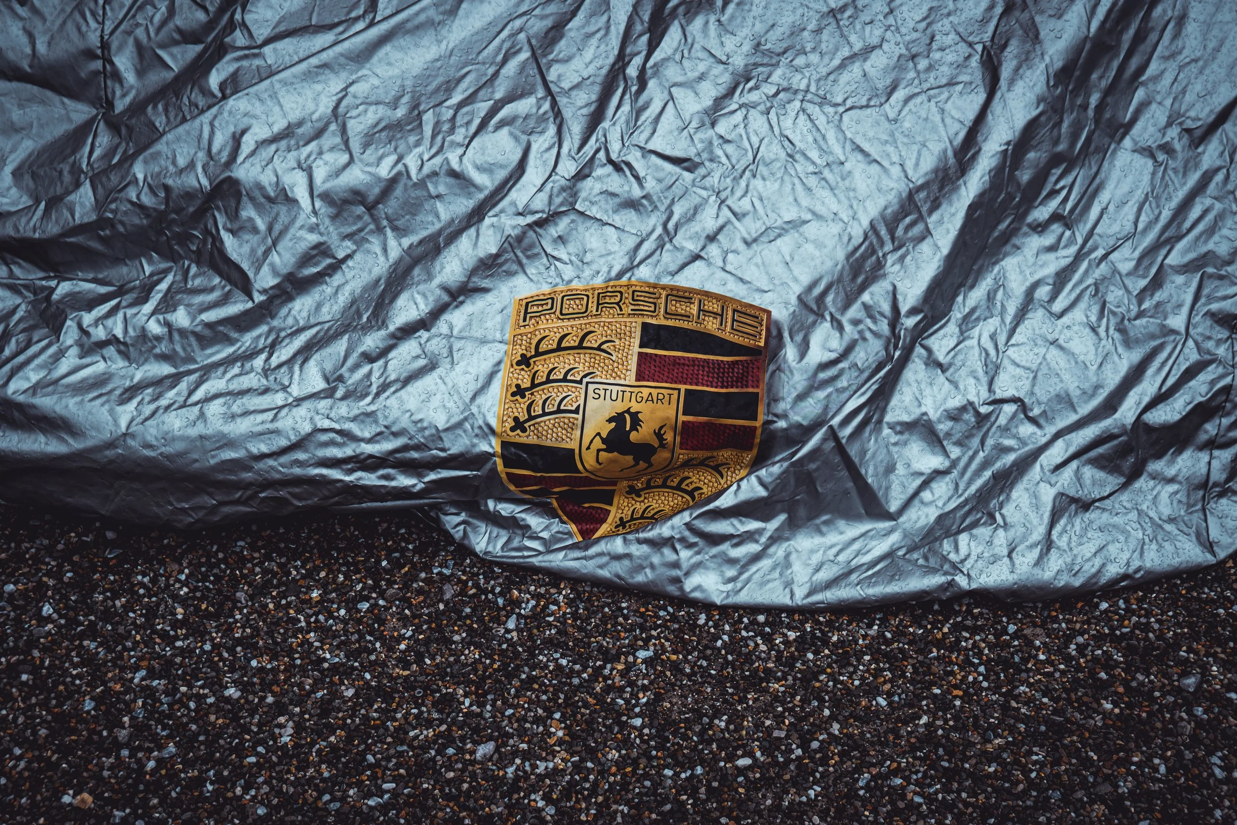 A crumpled gray material with raindrops on it, featuring a Porsche emblem for Stuttgart location in the lower center of the image.