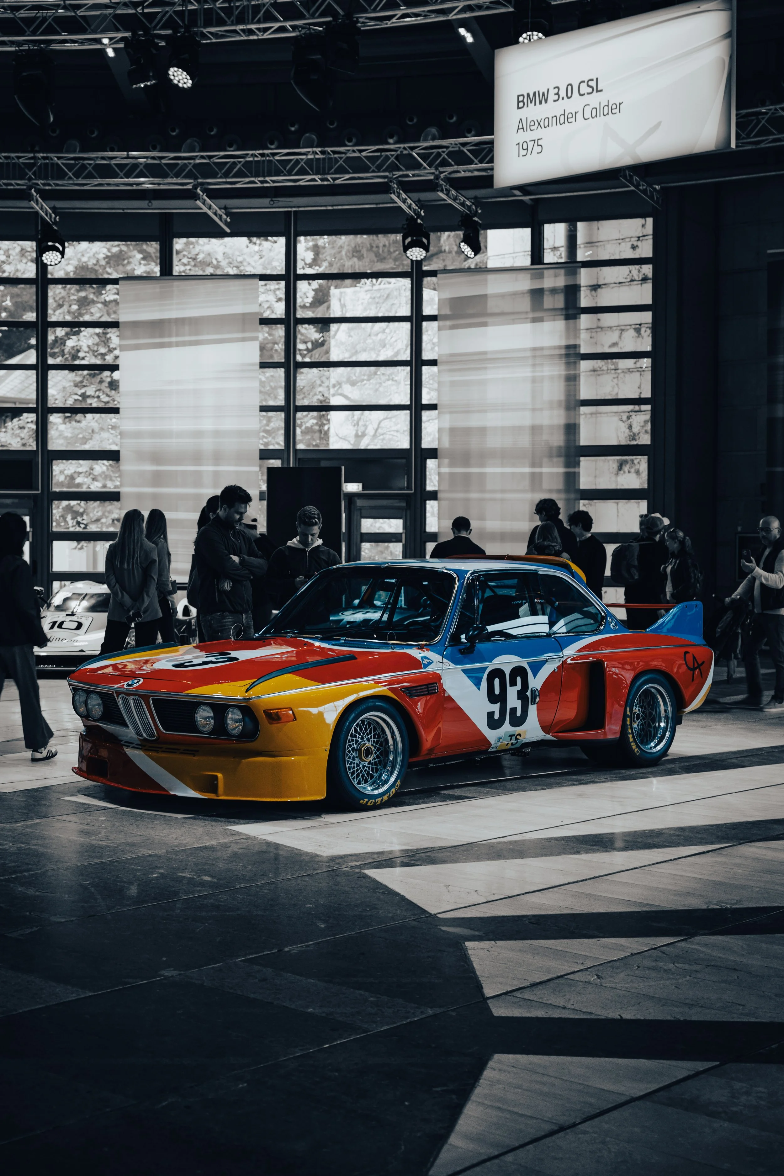 A vintage BMW race car with the number 93 on the side, painted in red, yellow, and blue, on display at an indoor automotive exhibit with people observing around.