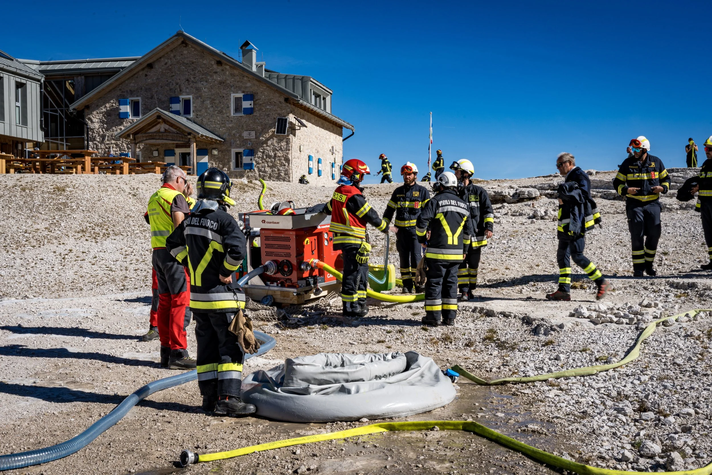 Firefighters and rescue personnel in protective gear working on a scene with a boat and equipment outside a stone house on a clear, sunny day.