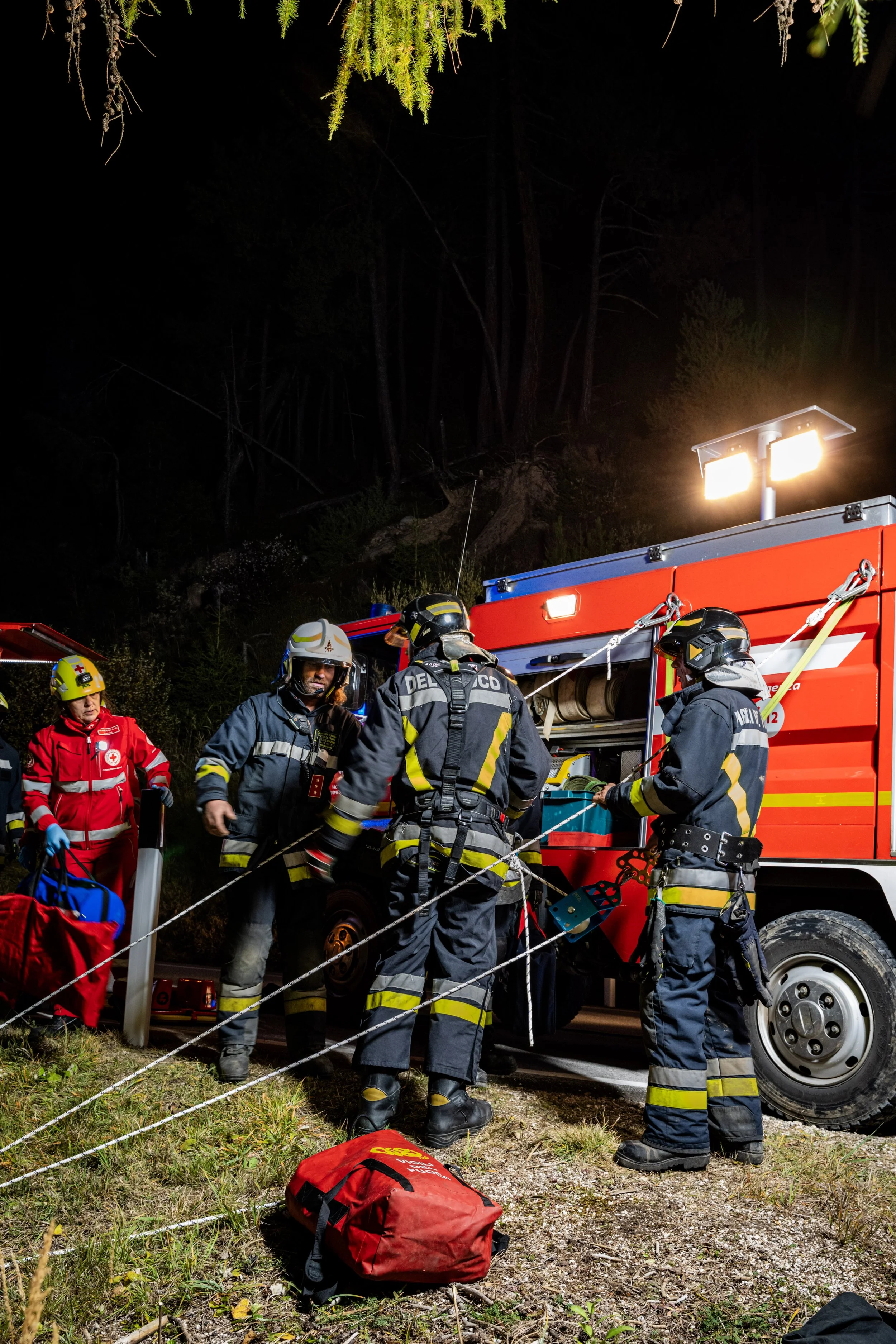 Firefighters and emergency personnel at night working near a fire truck with bright lights, with trees in the background and equipment on the ground.