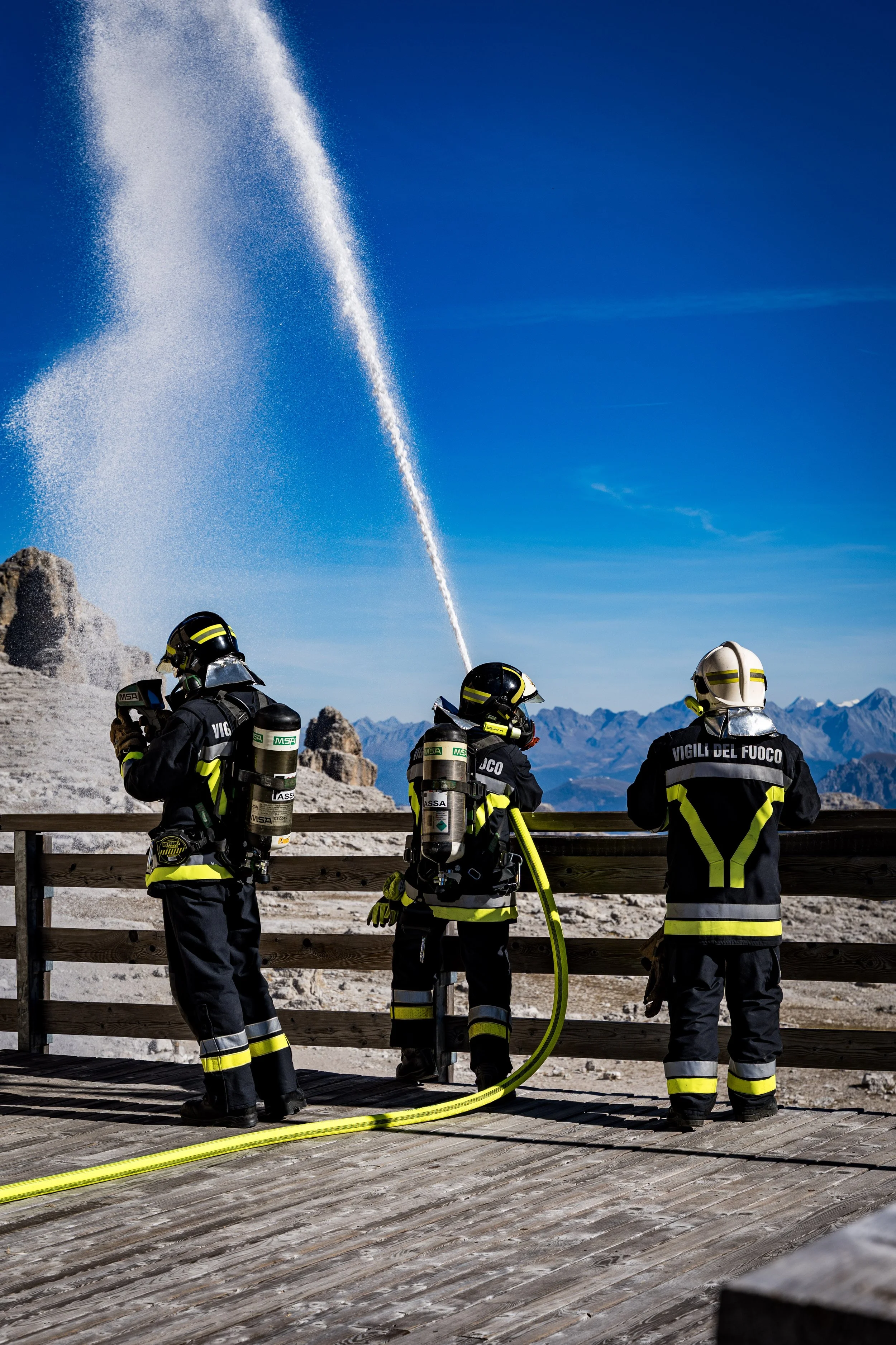 Firefighters in uniform and helmets standing on a wooden platform, handling a yellow fire hose amid a mountainous landscape under a clear blue sky with a water jet in the background.