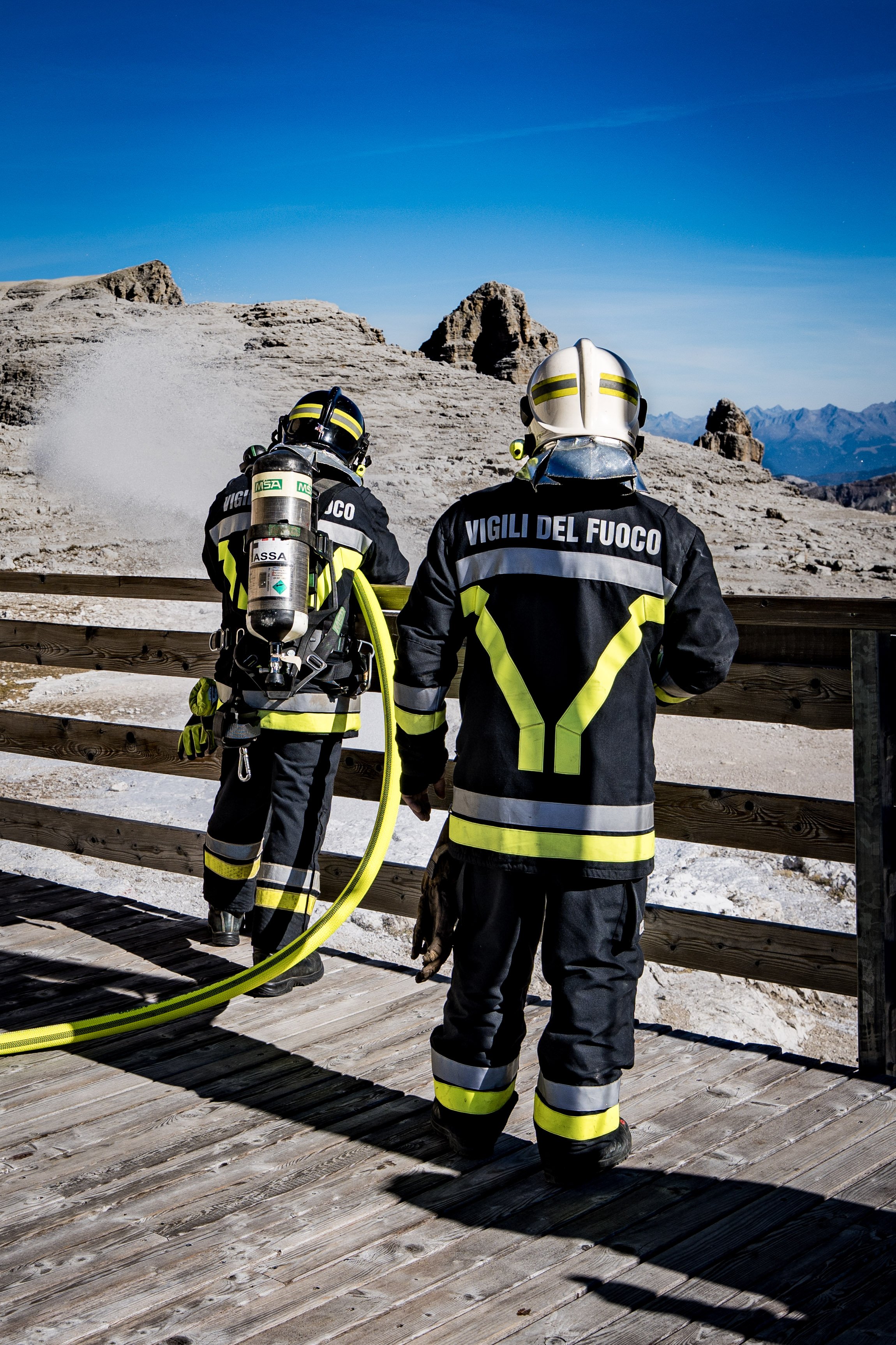 Two firefighters in protective gear, with one holding a yellow fire hose, stand on a wooden deck overlooking a landscape with rocky mountains and a clear blue sky.