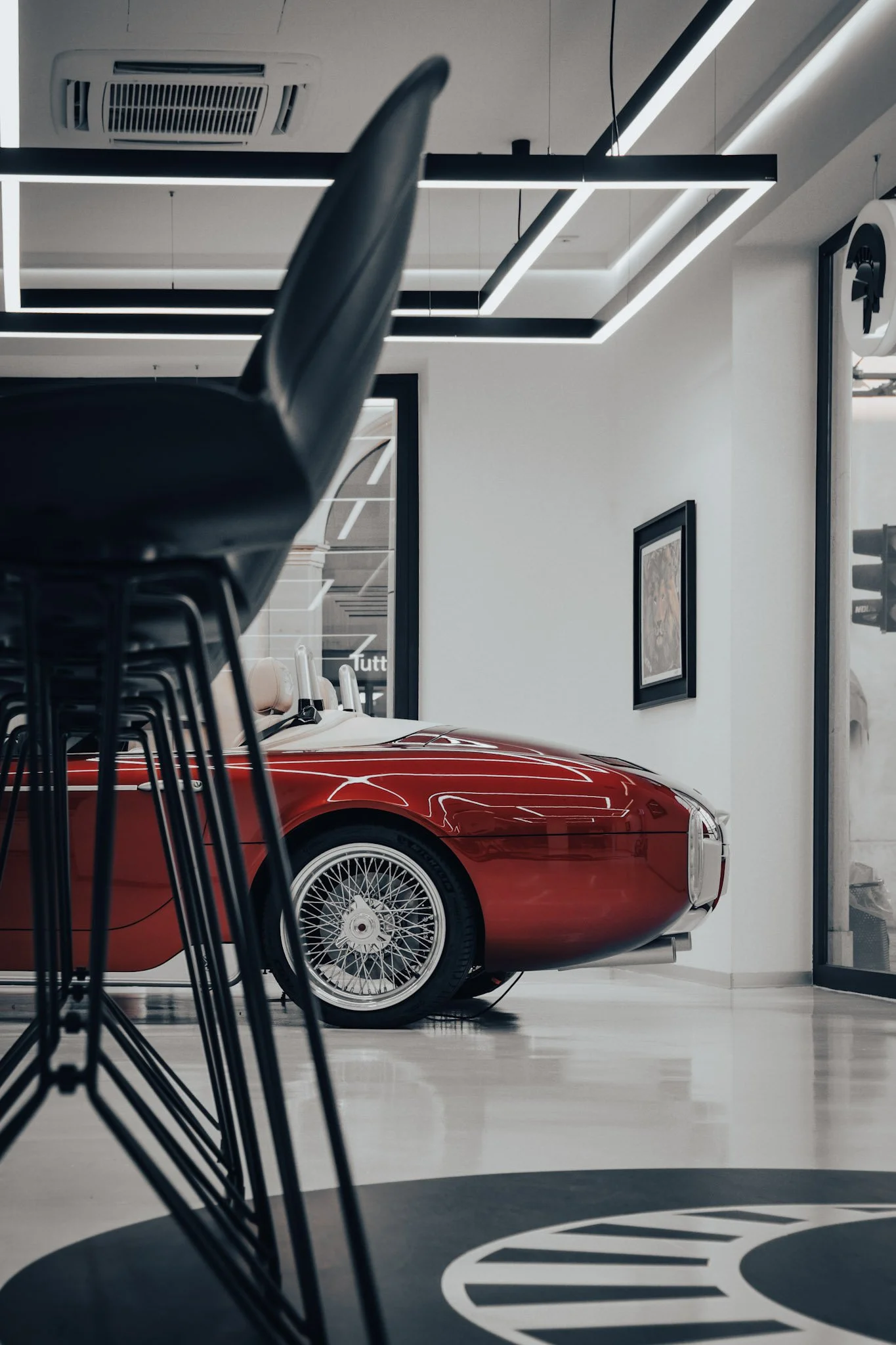 Red vintage car with wire-spoke wheels inside a modern showroom or garage, partially obscured by black chairs in the foreground, with framed artworks on white walls and a ceiling with linear LED lighting.