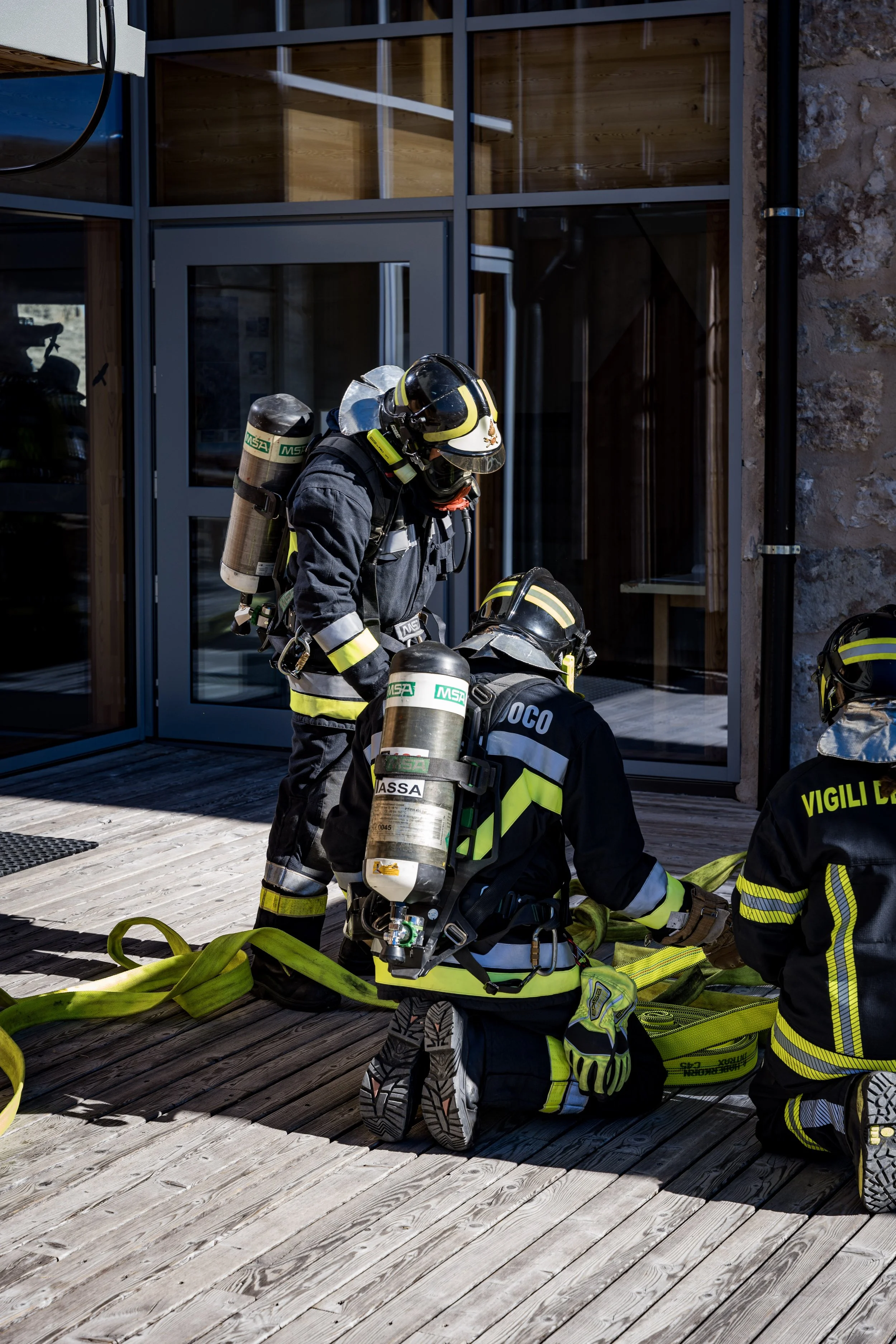 Firefighters in protective gear preparing hoses outside a building with large glass windows and stone wall.