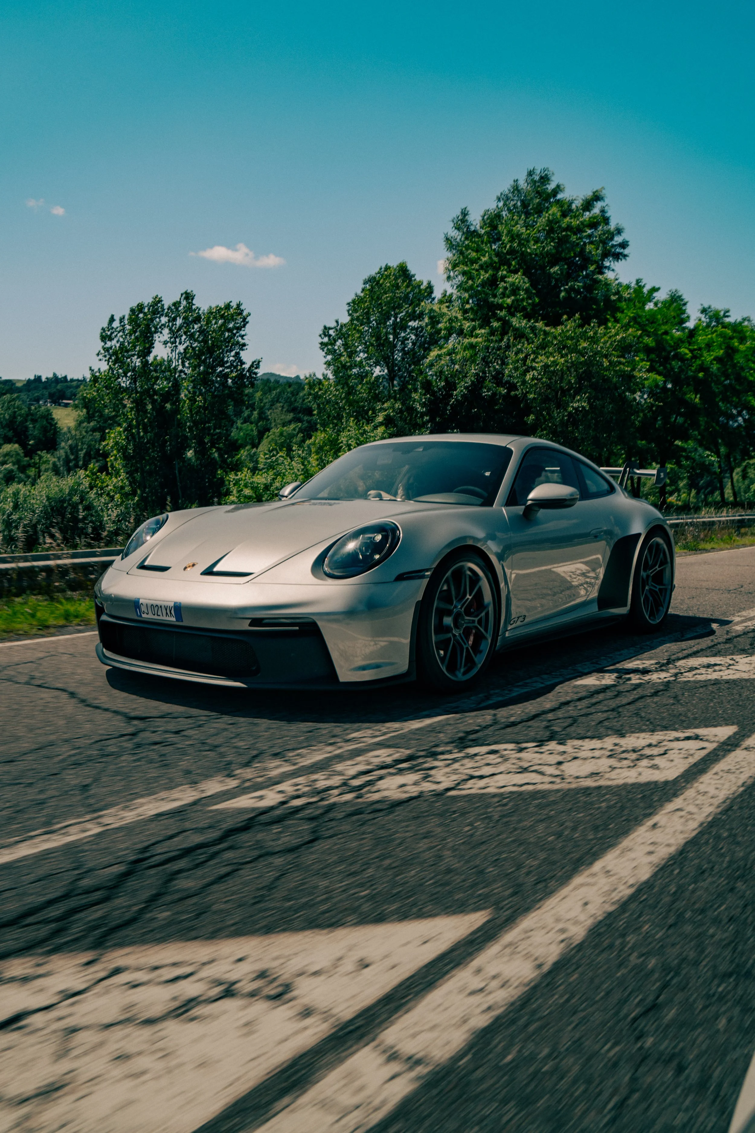 A silver Porsche sports car driving on a paved road with cracks, alongside a guardrail and lush green trees under a clear blue sky.