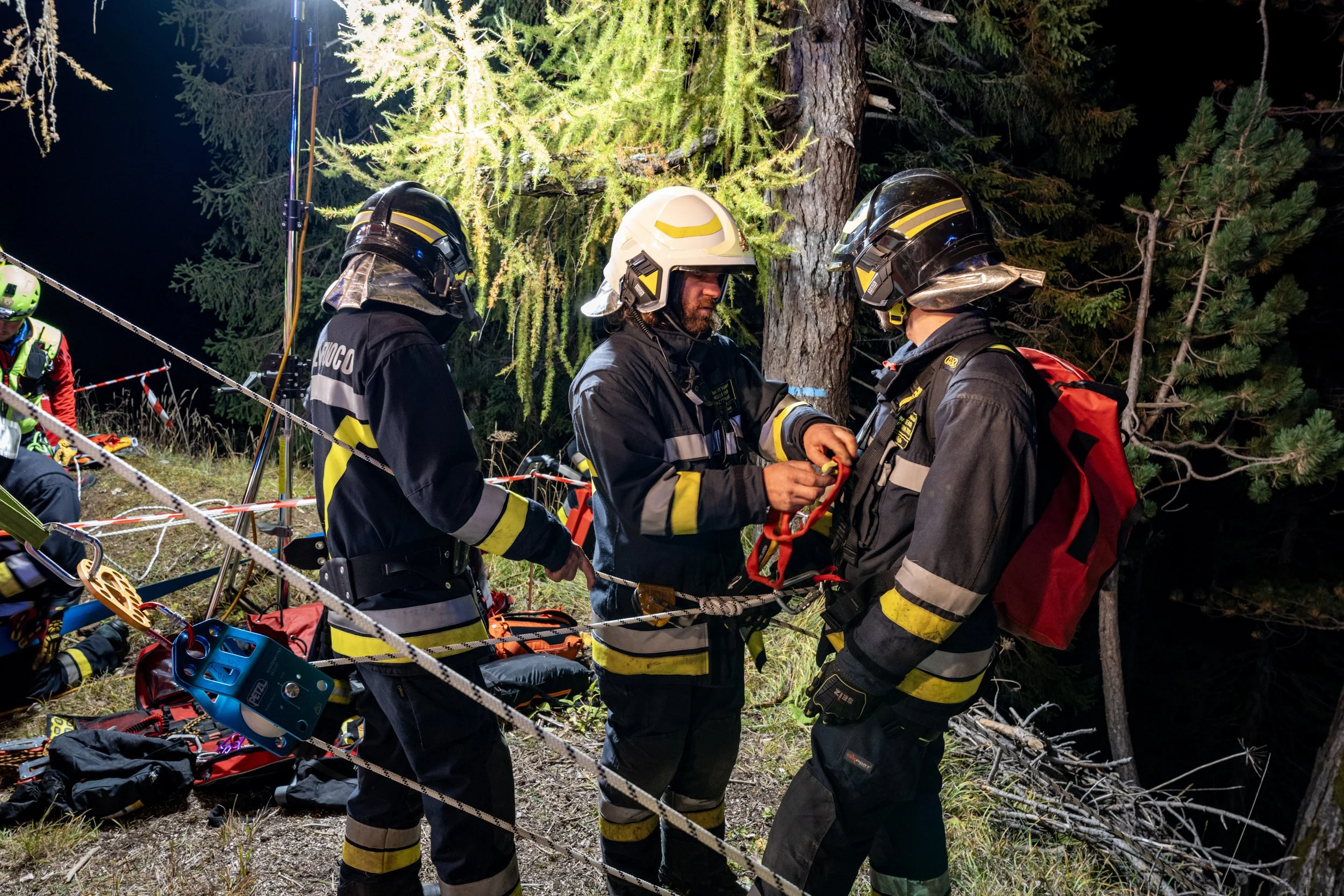 Nighttime rescue scene shows firefighters in protective gear working in a forested area with equipment. They are near a large tree with green foliage, surrounded by trees. One firefighter with a white helmet is talking to two other firefighters with 