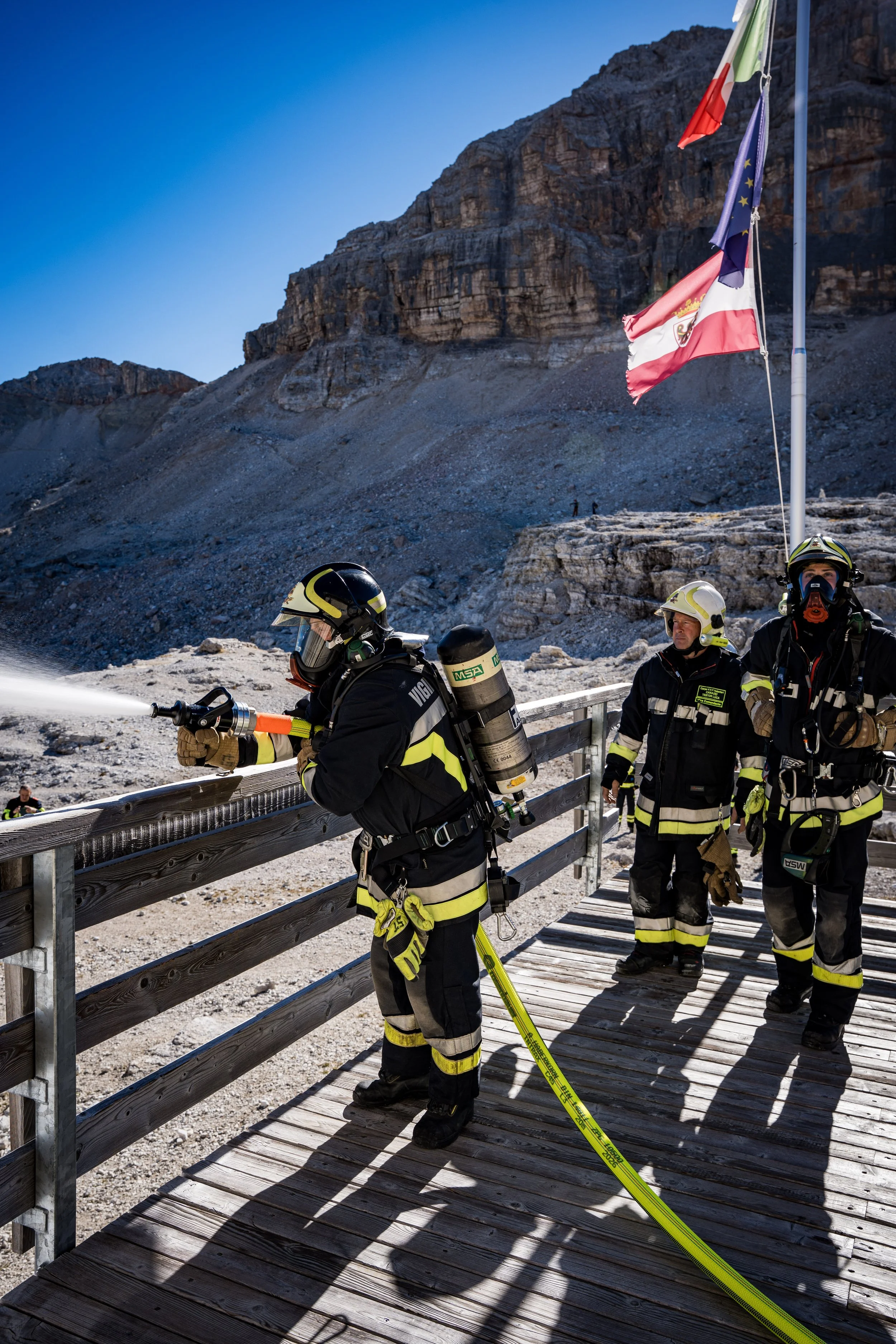 Firefighters dressed in firefighting gear and helmets stand on a wooden bridge, with a mountain and clear blue sky in the background. One firefighter is spraying water, and there are flags on poles nearby.