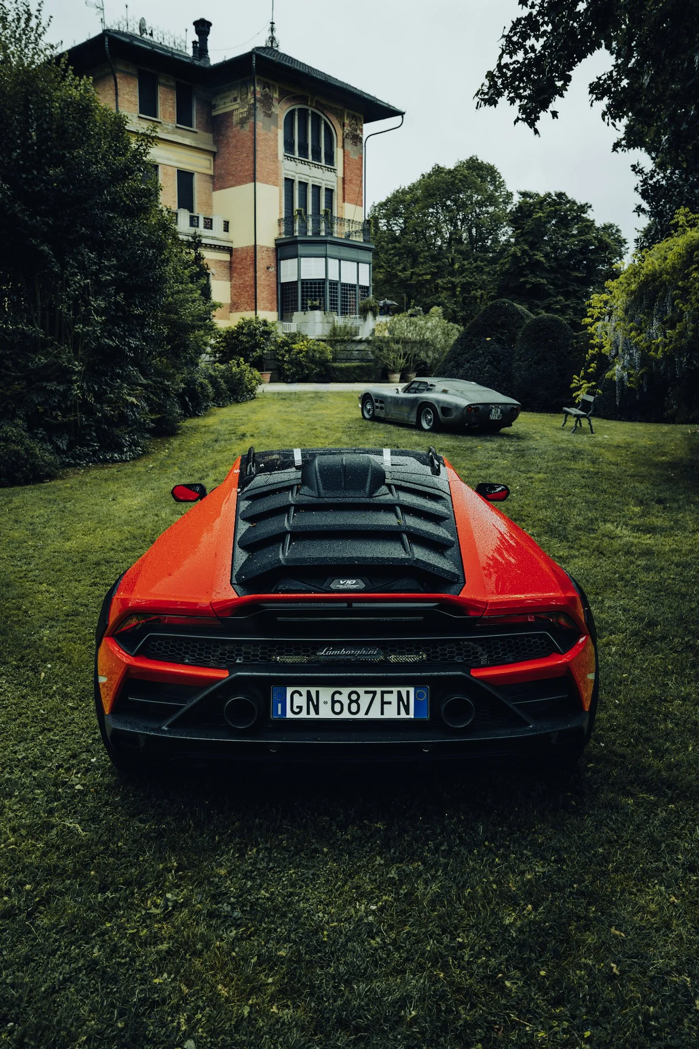 Red Lamborghini sports car with a European license plate, viewed from behind, parked on a grassy lawn. In the background, there are two vintage cars, one gray and one dark, near a large house with a brick and cream facade surrounded by greenery, tree