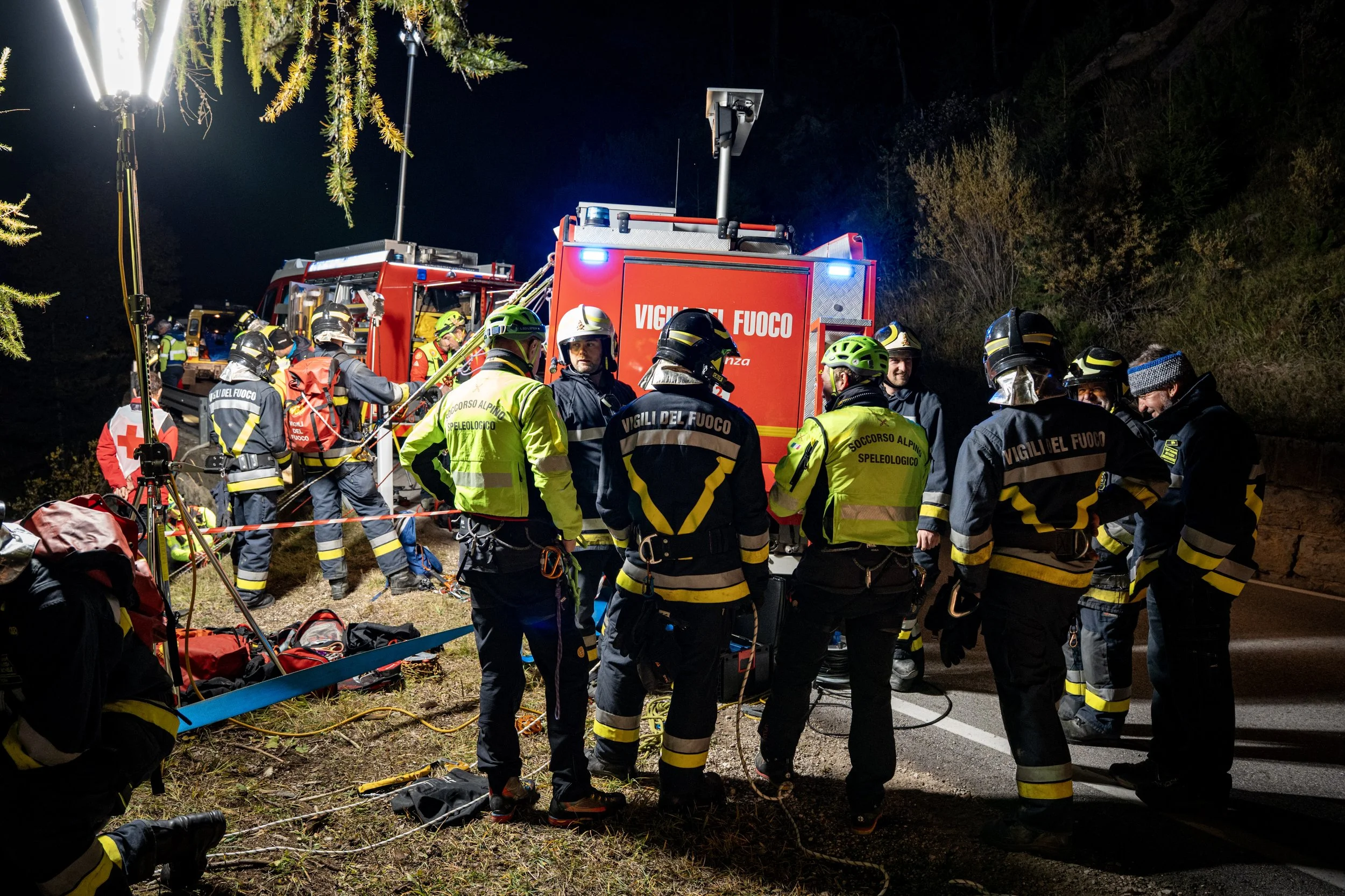 Nighttime scene of emergency responders, including firefighters, gathered around a fire truck with flashing lights on a road. They are preparing gear and equipment, possibly at the scene of an accident or rescue operation.
