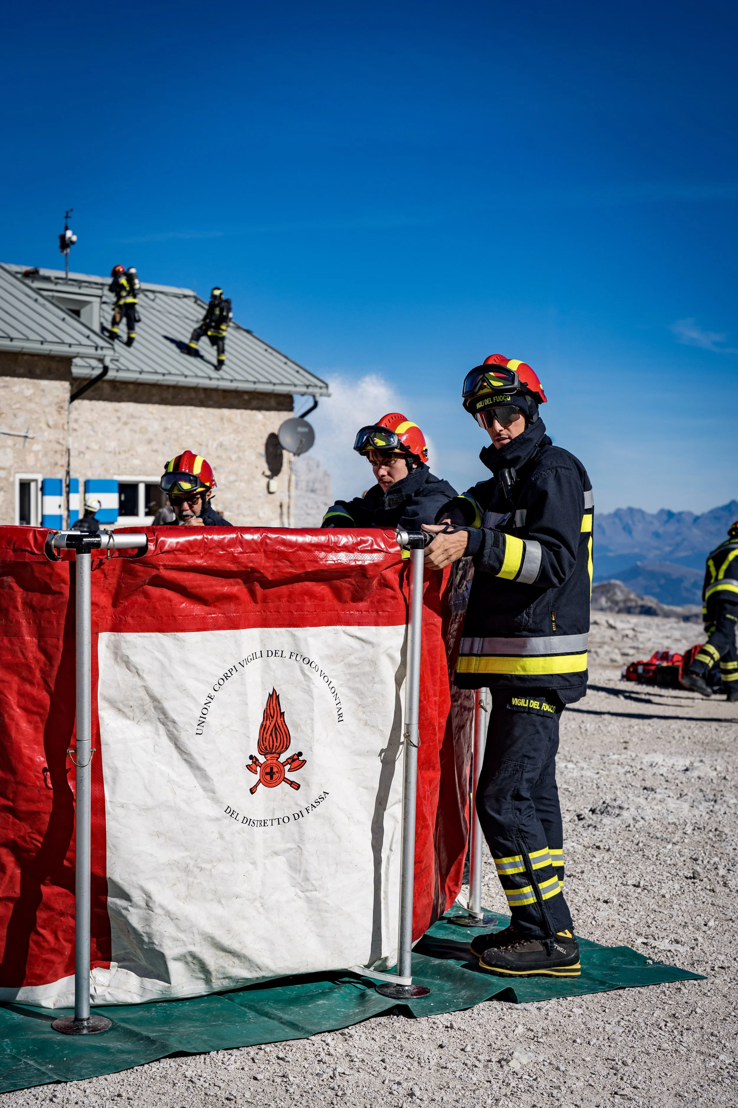Firefighters in protective gear and helmets standing behind a red and white emergency barrier with Italian text and emblem, with some firefighters on a roof and mountains in the background under clear blue sky.
