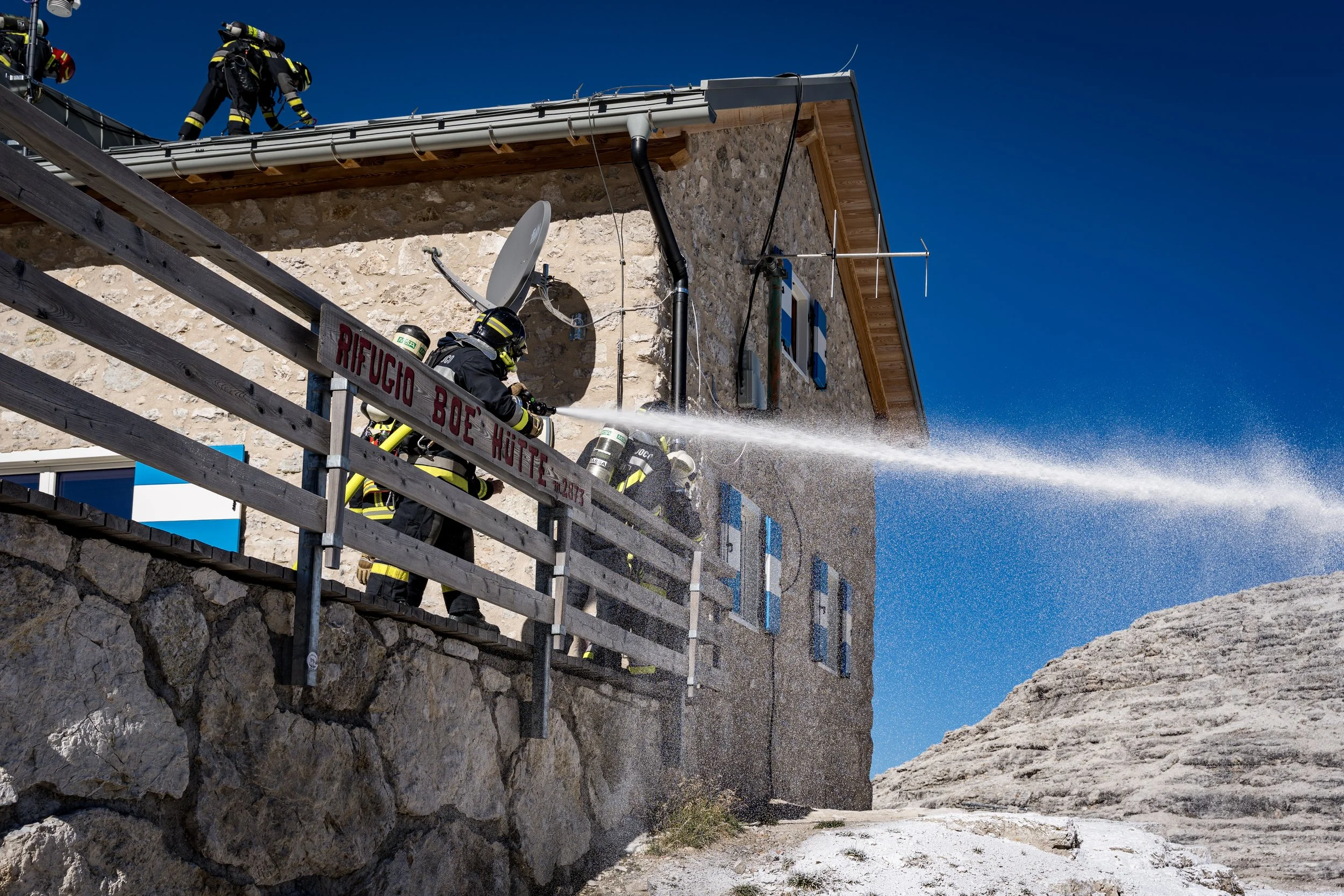 Firefighters spraying water from a hose on a stone building with a wooden roof. Two firefighters are on a balcony, working to extinguish a fire. The building has blue window shutters, a satellite dish, and an antenna on the roof. The sky is clear and