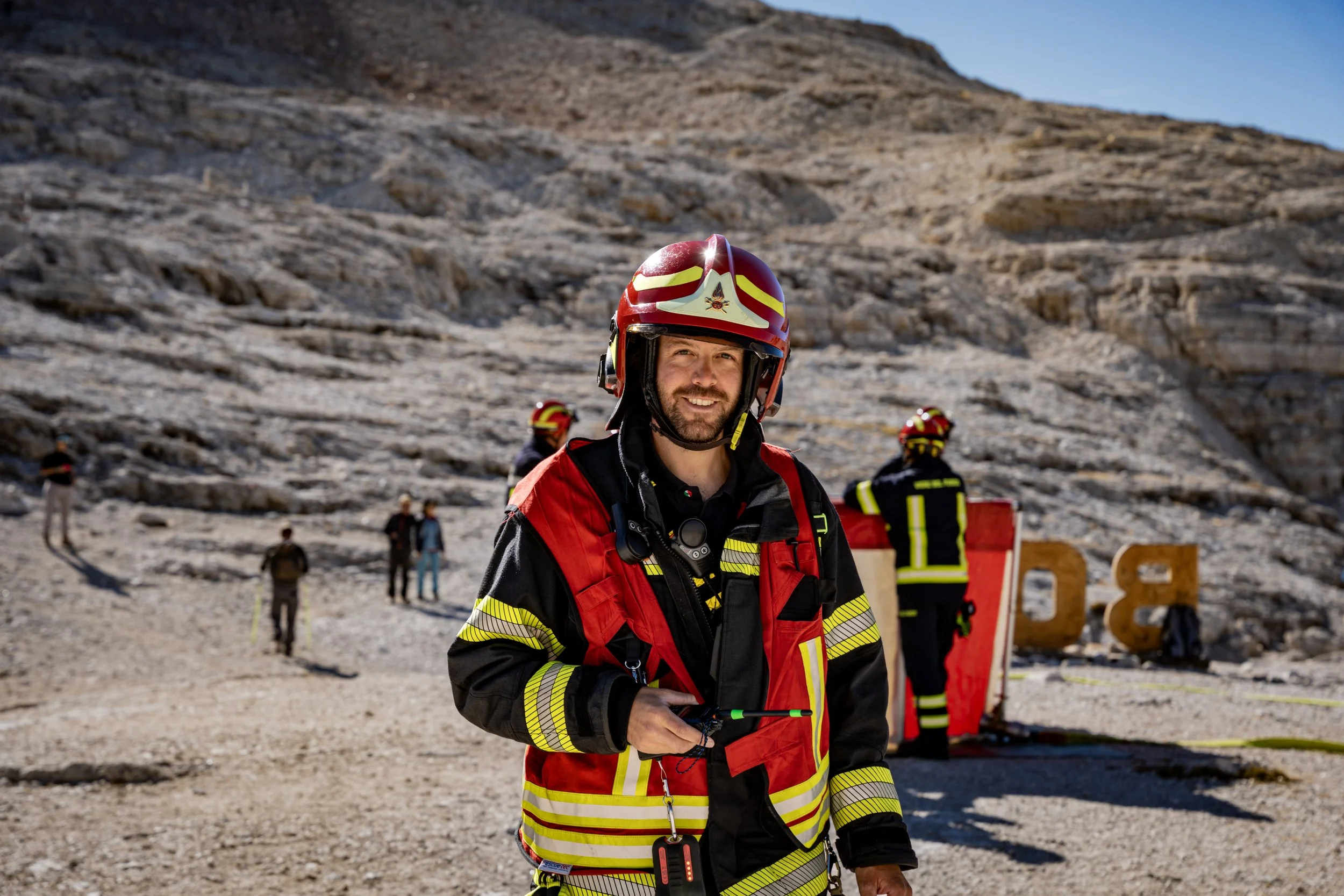 A smiling firefighter in red and black gear with yellow reflective stripes, wearing a red helmet with yellow markings, standing outdoors at a rocky, mountainous area with other crew members and safety equipment in the background.
