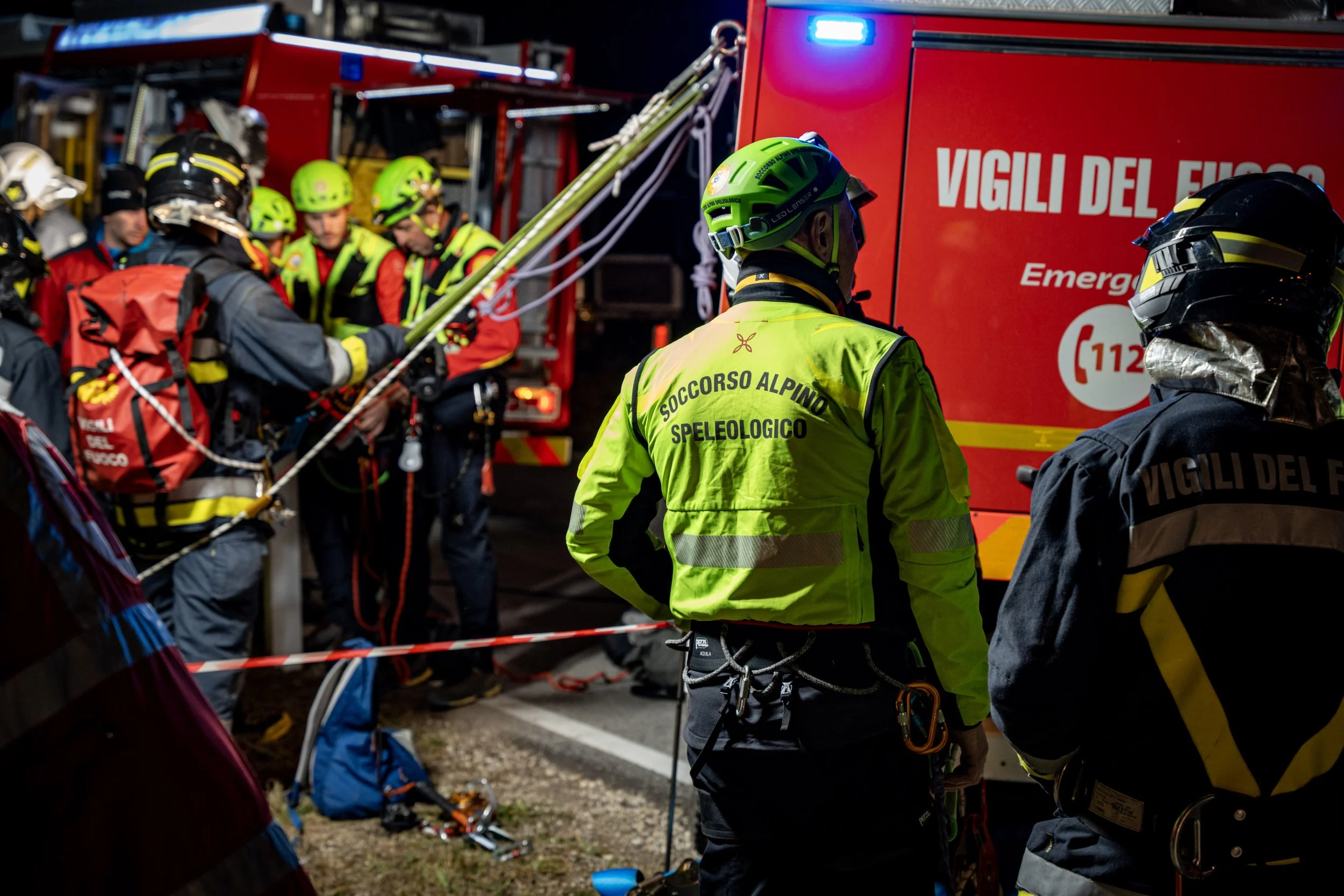 Rescue team members wearing helmets and high-visibility jackets working near an emergency vehicle marked 'VIGILI DEL FUOCO.'