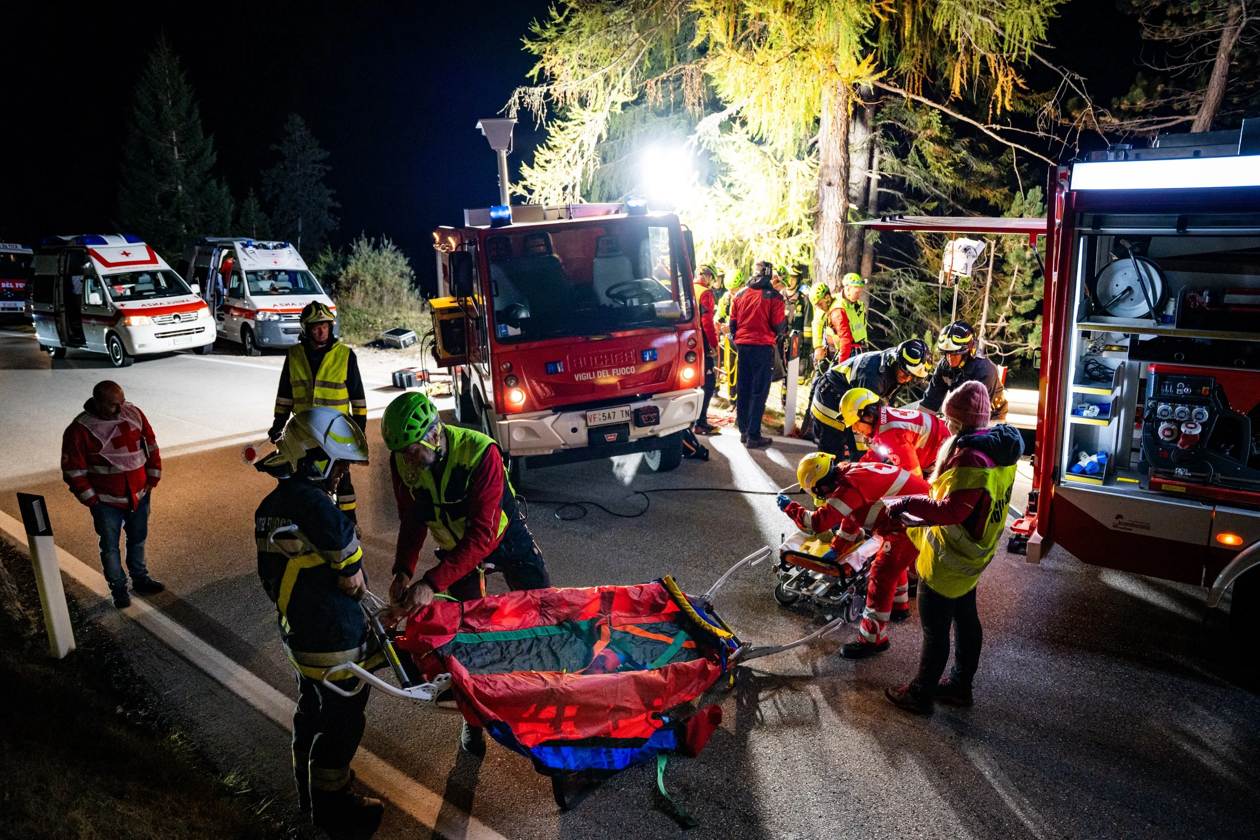 Night scene of emergency responders, firefighters, and paramedics attending to an incident in a wooded area, with emergency vehicles and equipment on the road.