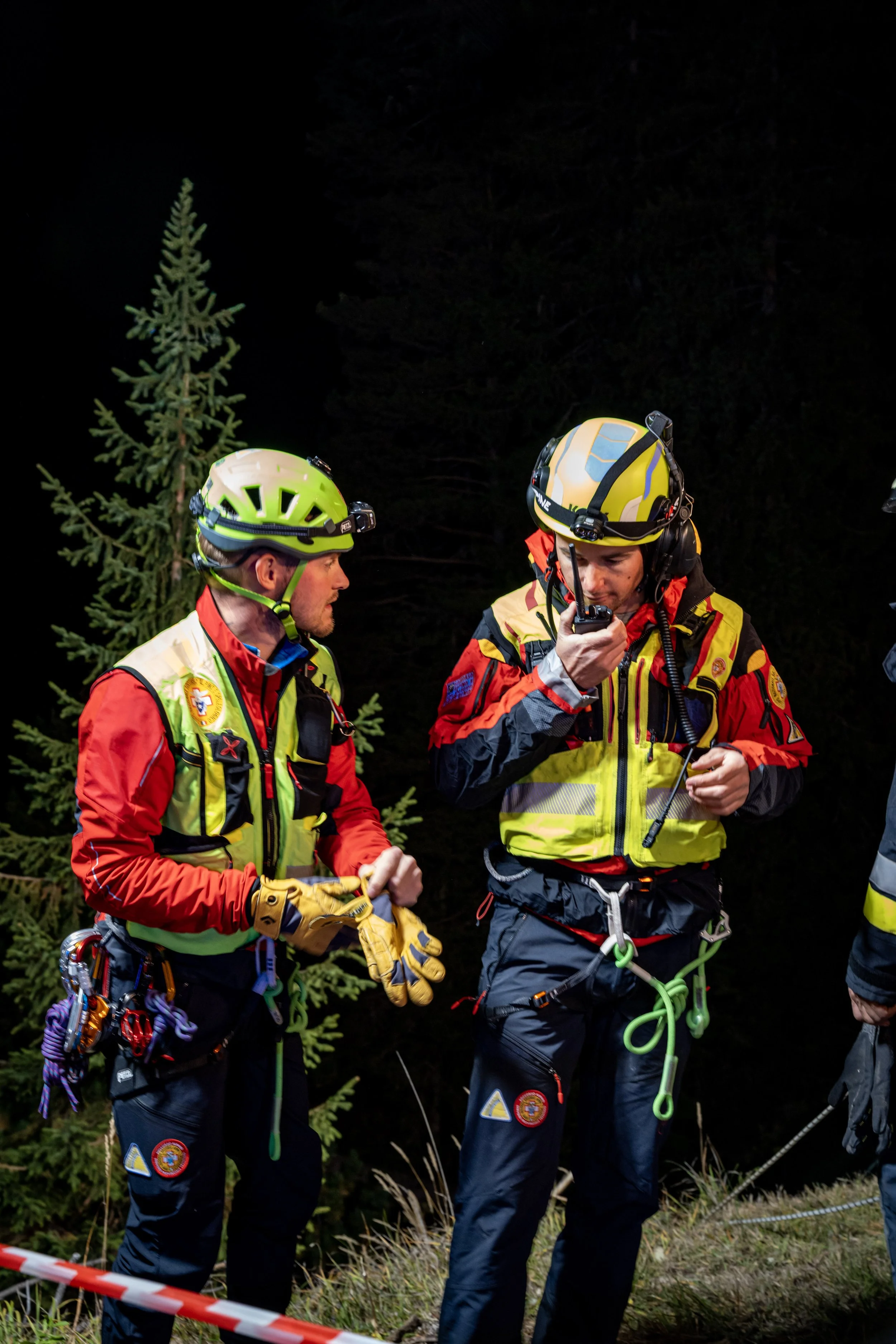 Two rescue workers in bright jacket and helmet talking at night in a forested area, with trees and grass around, one holding a walkie-talkie.