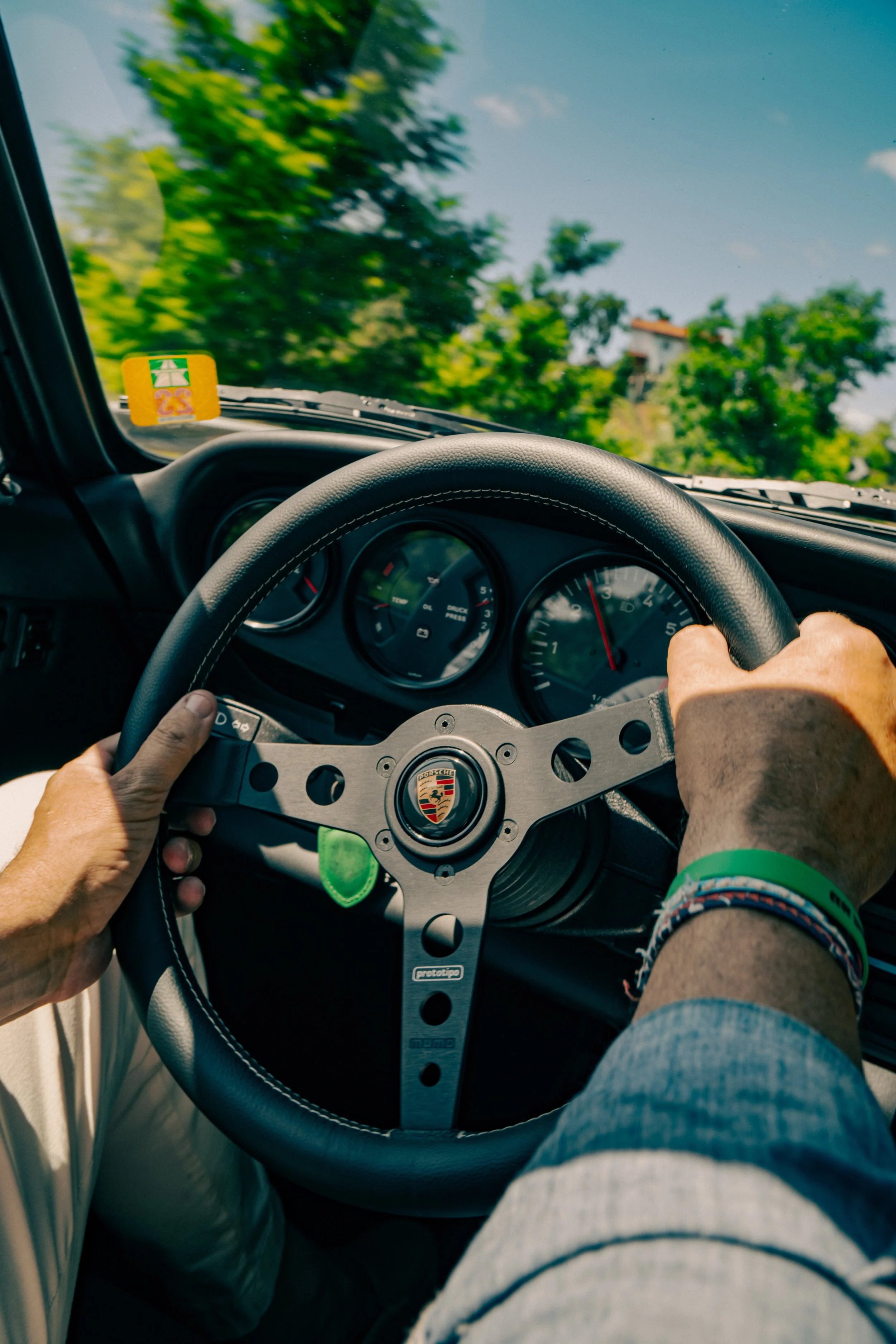 A first person view of a person driving a Porsche car with hands on a black steering wheel, speedometer, and a dashboard. Outside, trees and a blue sky are visible through the windshield.