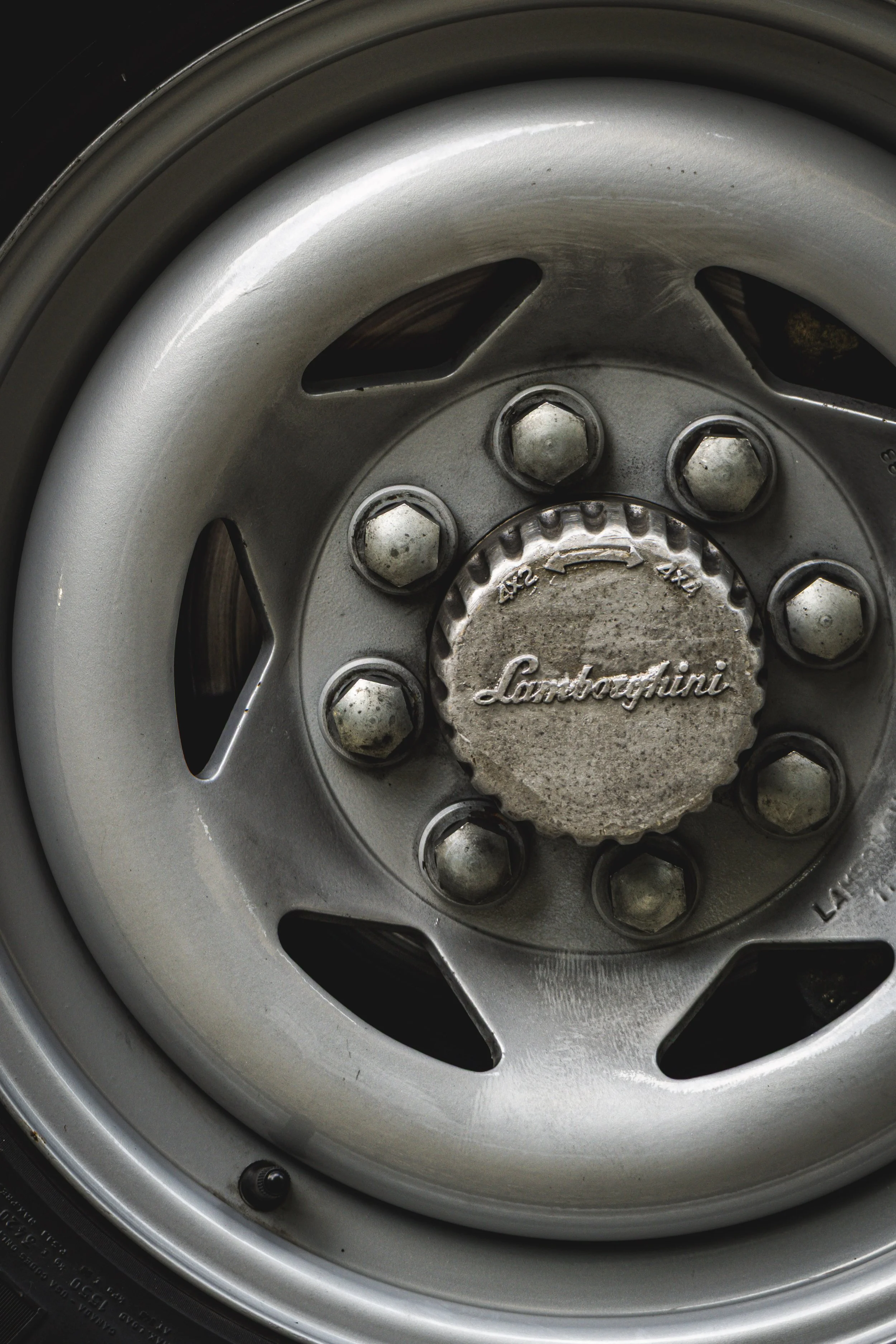 Close-up of a silver wheel rim with a center cap that has the Lamborghini logo engraved, surrounded by lug nuts and wheel spacers, with a black tire partially visible.