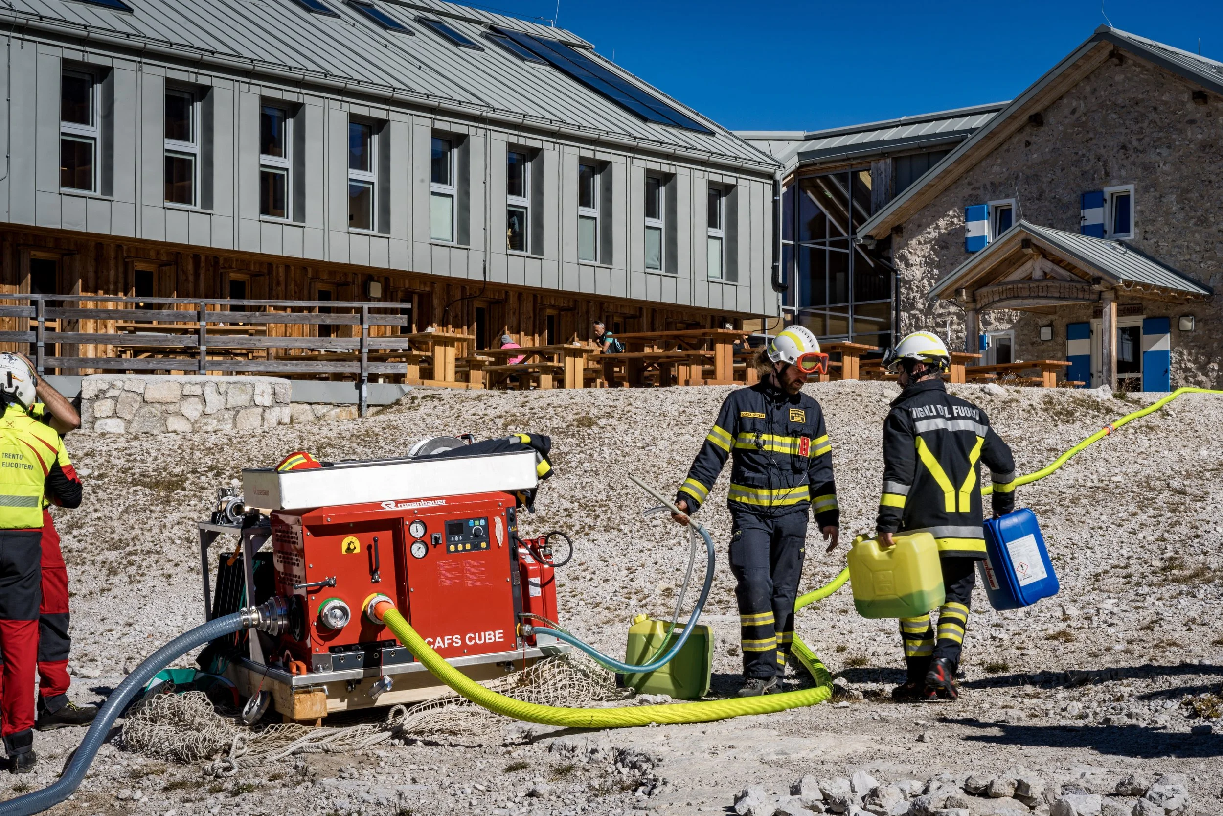 Firefighters and rescue personnel in safety gear working with water hoses and firefighting equipment outside a building under construction or renovation.