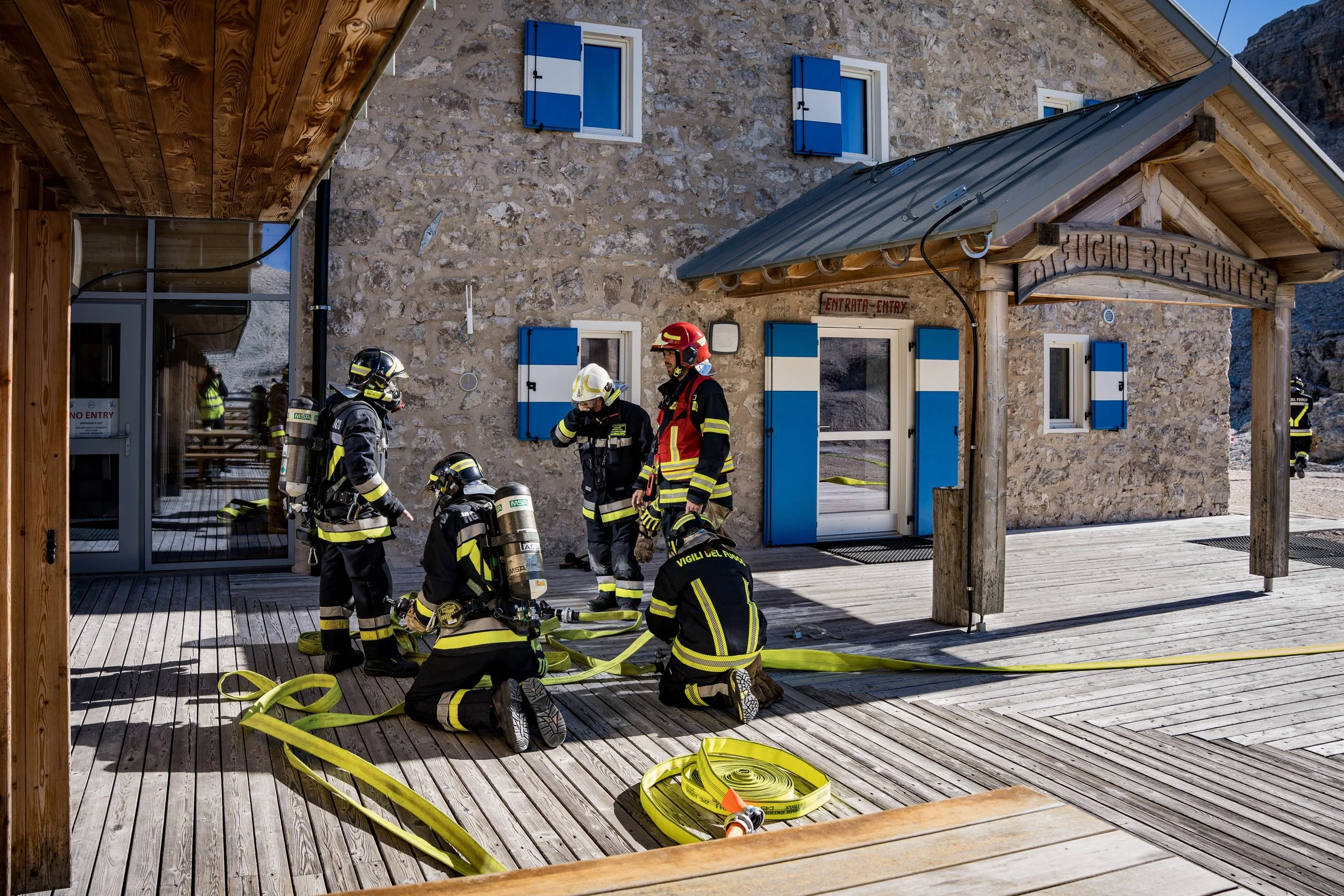 Firefighters in protective gear preparing equipment outside a stone building with blue shutters and a sign that reads "FUGIO 3.5".